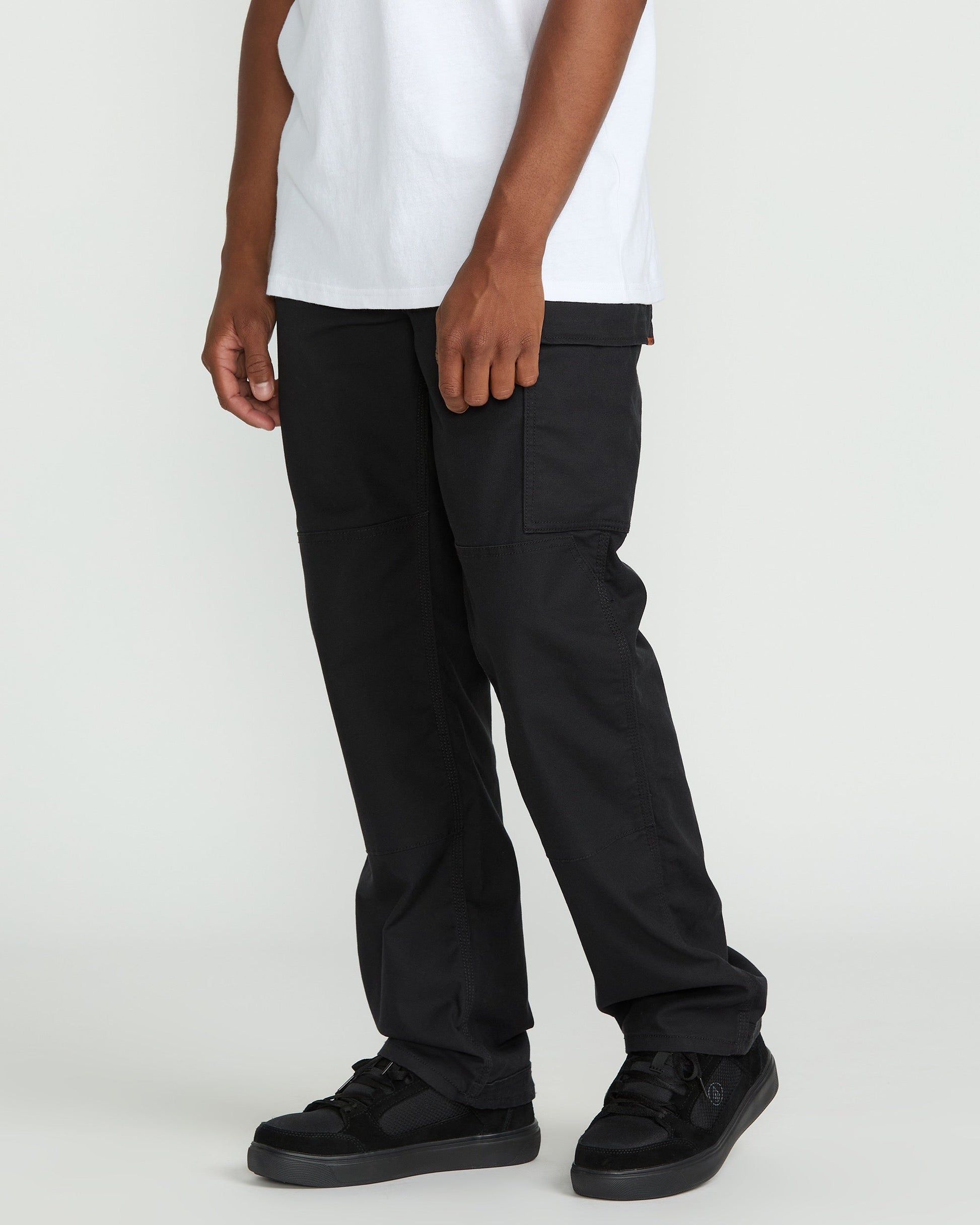 A person, visible only from the waist down, stands against a plain light background wearing Volcom Men's Caliper II Relaxed Work Pants with cargo pockets, paired with a white t-shirt and black sneakers.
