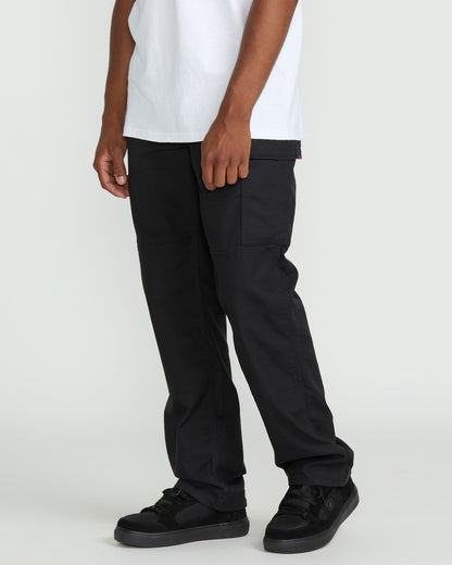 A person, visible only from the waist down, stands against a plain light background wearing Volcom Men's Caliper II Relaxed Work Pants with cargo pockets, paired with a white t-shirt and black sneakers.