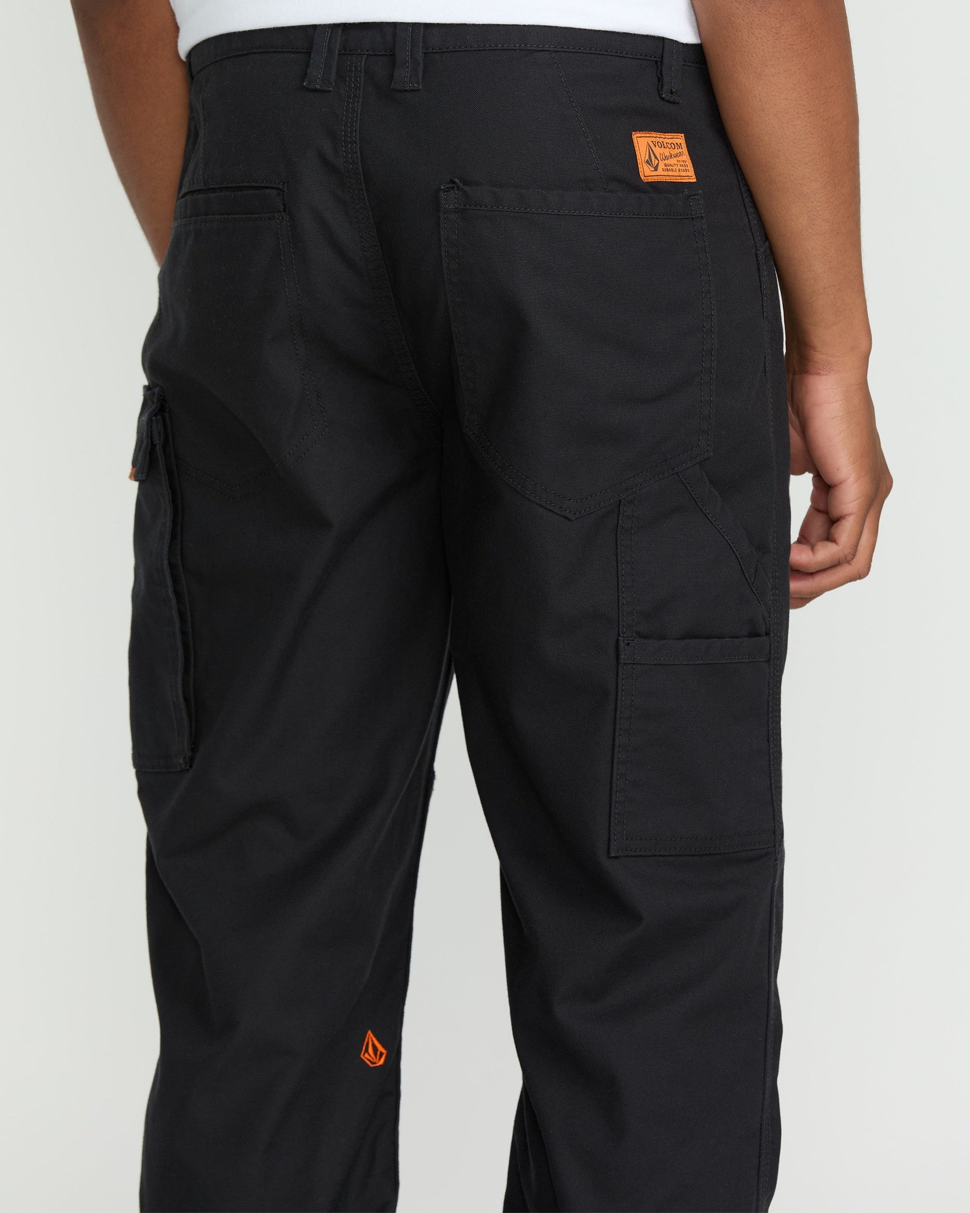 A person wears Volcom Men's Caliper II Relaxed Work Pants in black stretch canvas with multiple cargo pockets and orange logo patches, paired with a white shirt. The image shows the back view, highlighting pocket and stitching details.