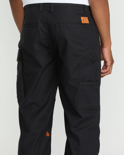 A person wears Volcom Men's Caliper II Relaxed Work Pants in black stretch canvas with multiple cargo pockets and orange logo patches, paired with a white shirt. The image shows the back view, highlighting pocket and stitching details.