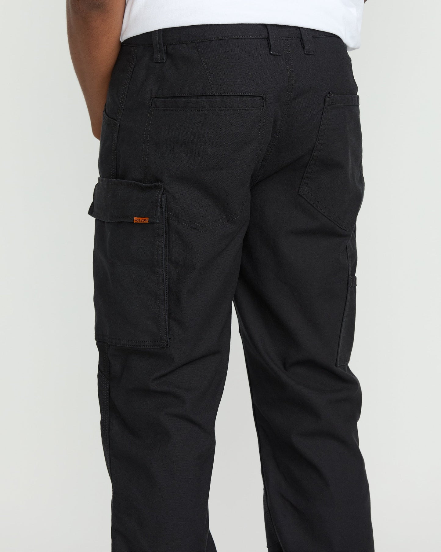 A person wearing Volcom Men's Caliper II Relaxed Work Pants in black stretch canvas with multiple pockets, including a left-leg cargo pocket, and a white shirt; shown from the waist down and behind.