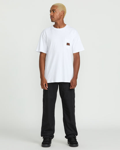 A person with short blond hair stands slightly to the side, wearing a white pocket t-shirt, Volcom Men's Caliper II Relaxed Work Pants in black stretch canvas with cargo pockets, and black sneakers against a plain white background.