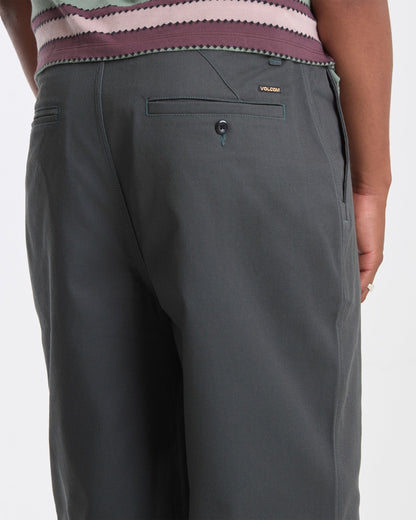 A person wearing Volcom Men's Billow Pants in dark gray stands with their back to the camera, revealing the back pocket, part of their hand, and the waistband of a patterned shirt.