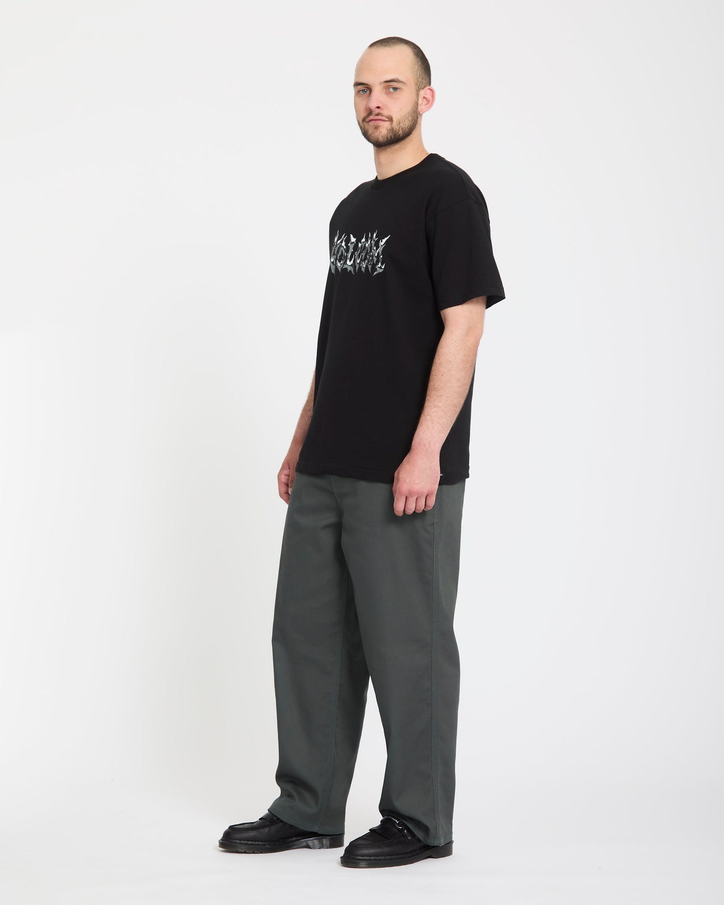 A bearded man stands against a plain white background, wearing a black graphic T-shirt, black shoes, and Volcom Men's Billow Pants in dark gray. He looks slightly to the side with a neutral expression.