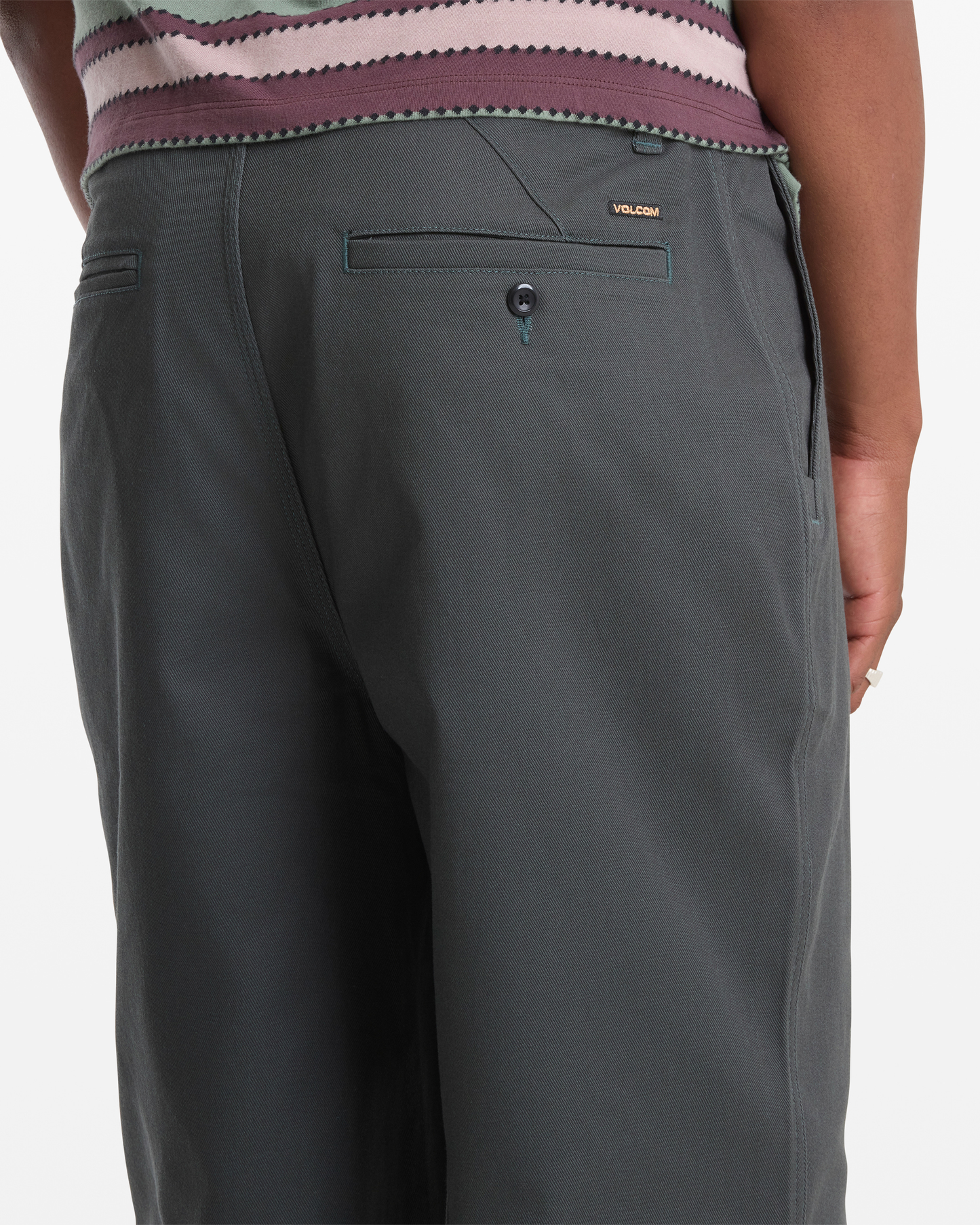 A person stands with their back to the camera, wearing Volcom Men's Billow Pants in dark gray. The pants feature a buttoned back pocket and a small gold Volcom label above the pocket.