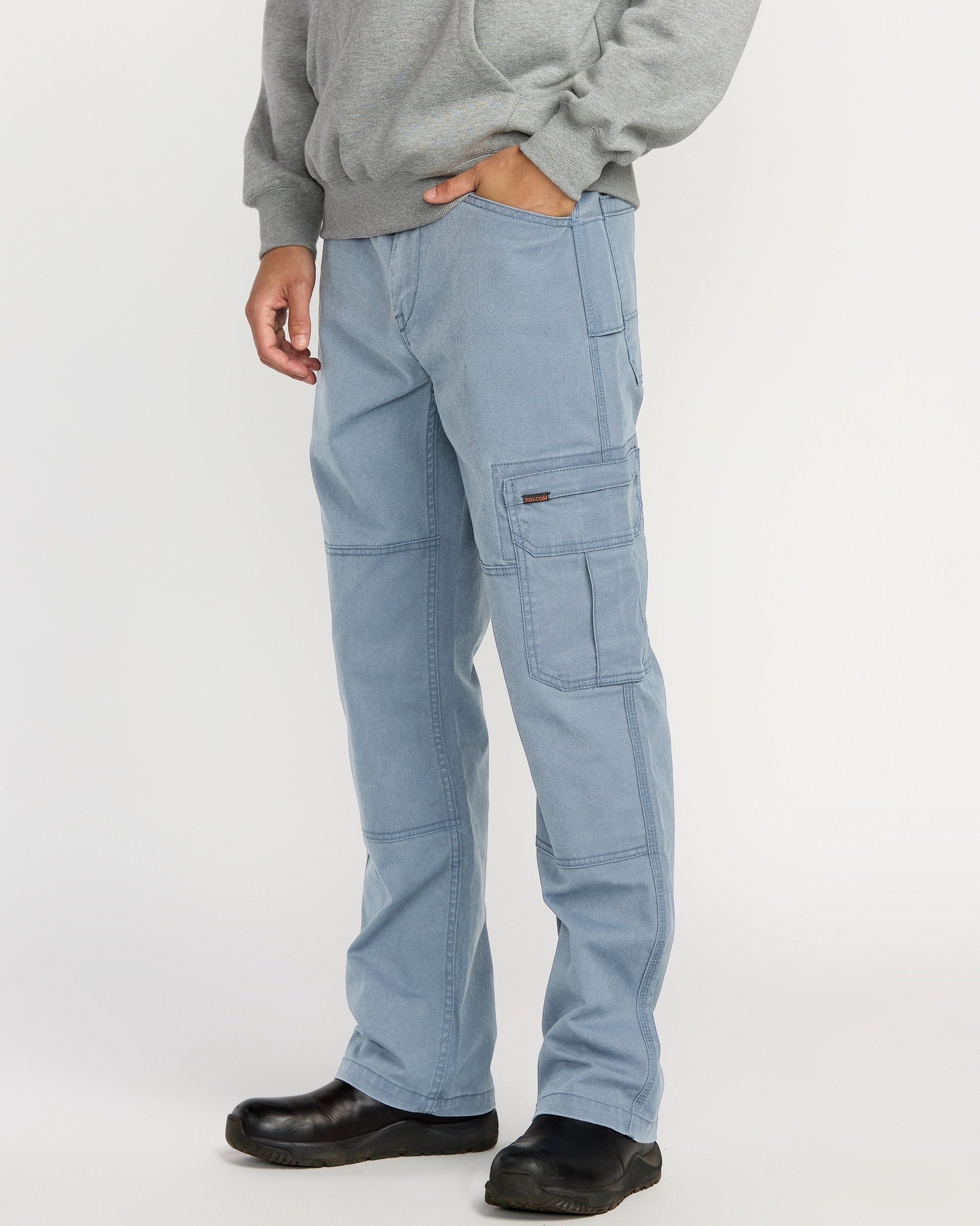A person models Volcom Men’s Gage Work Pants in light blue, paired with a gray hoodie and black shoes against a white background. One hand is in their pocket, and their face is not shown.