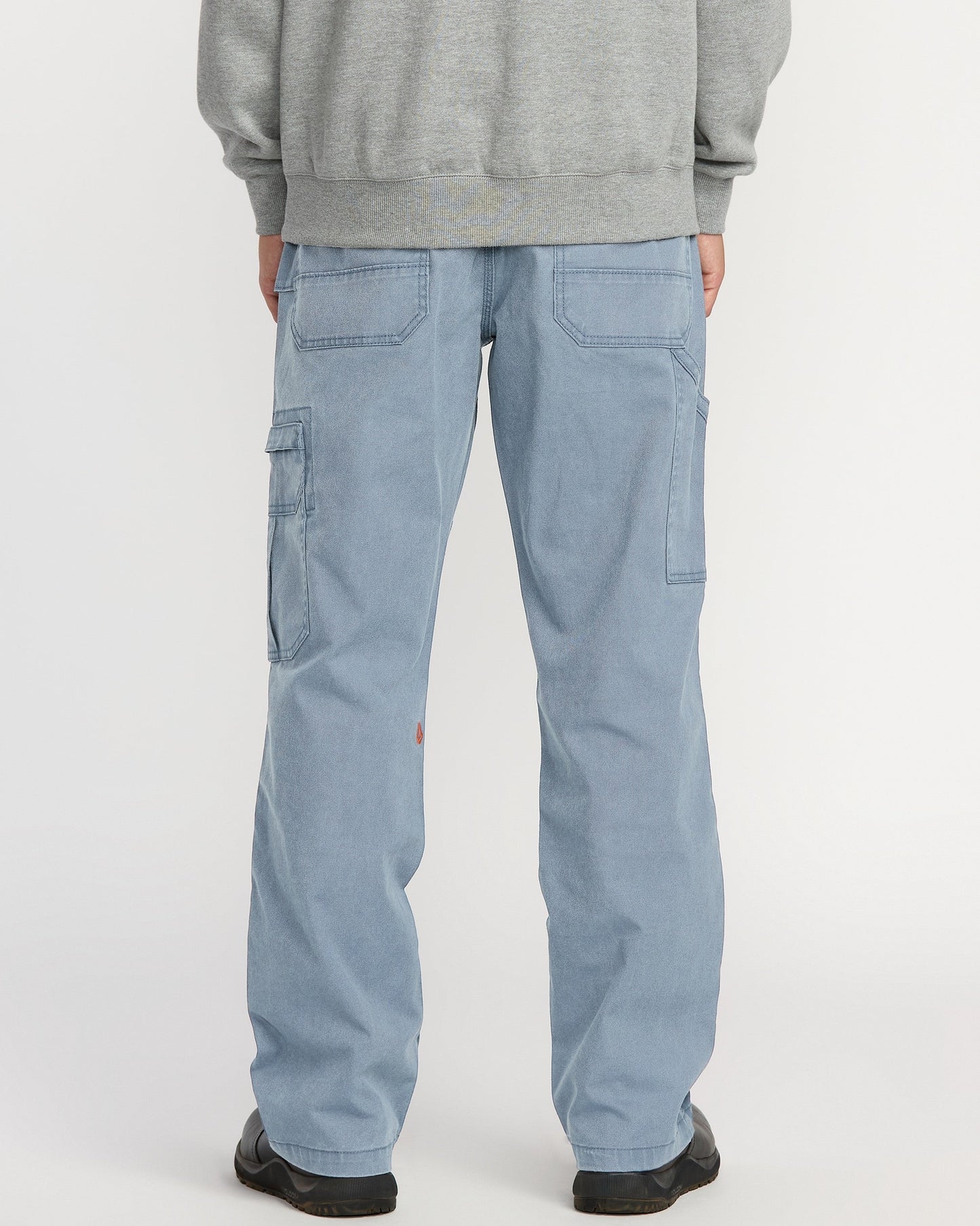 A person is shown from the back wearing Volcom Men's Gage Work Pants in light blue denim with large back and side pockets, paired with a gray sweatshirt, standing against a white background.