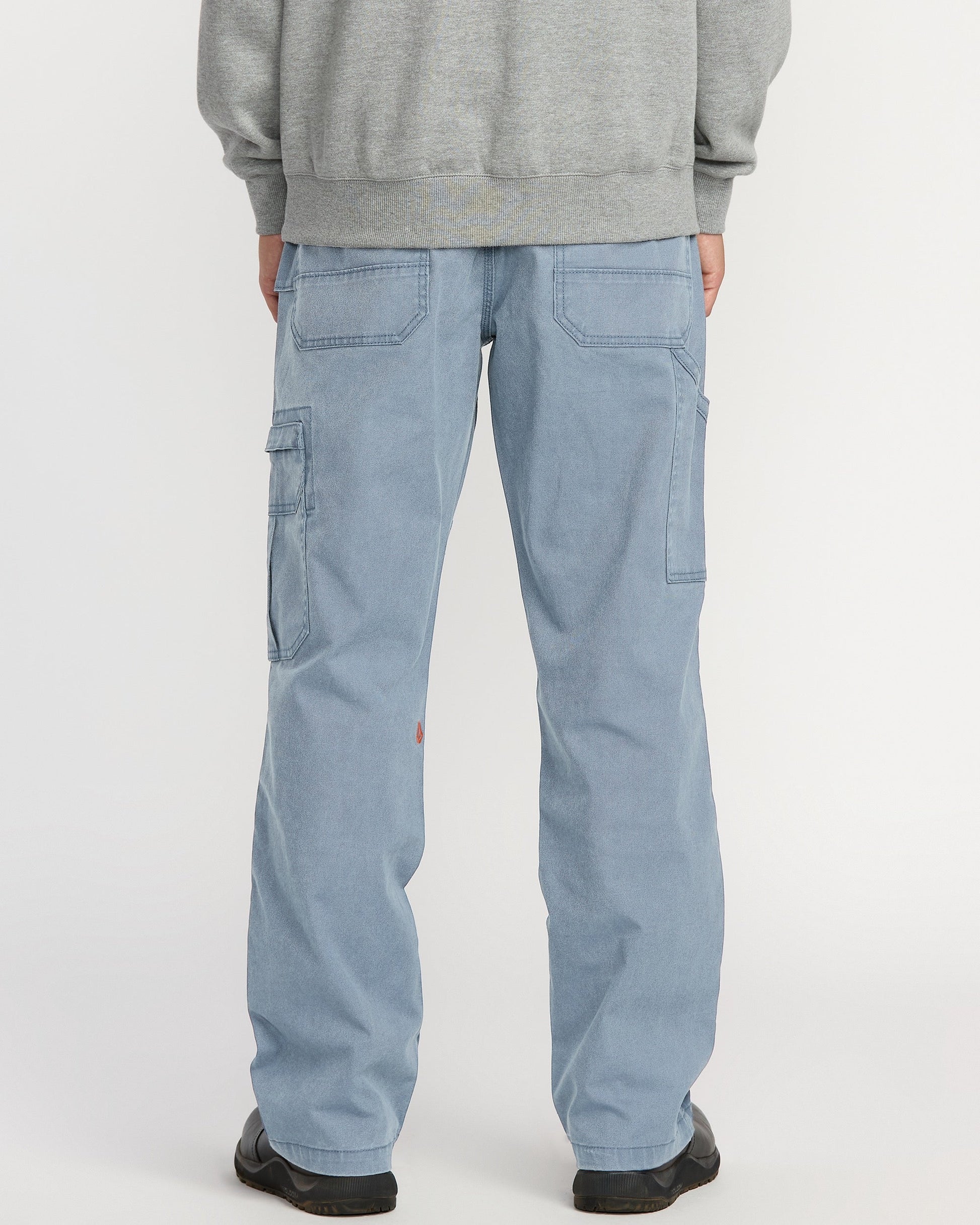 A person is shown from the back wearing Volcom Men's Gage Work Pants in light blue denim with large back and side pockets, paired with a gray sweatshirt, standing against a white background.