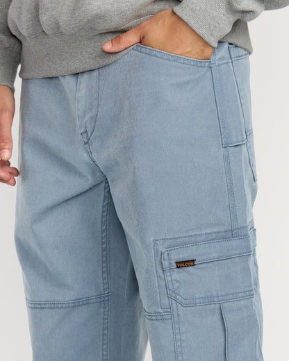 A person wears Volcom Men's Gage Work Pants in light blue, featuring visible stitching and a small logo tag on the side pocket, paired with a gray sweatshirt and one hand in their pocket.