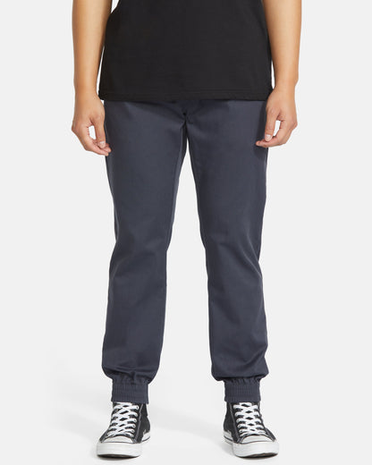 A person wearing a black t-shirt, Volcom Men's Frickin Slim Joggers in dark blue with elastic cuffs, and black-and-white sneakers stands against a plain white background.
