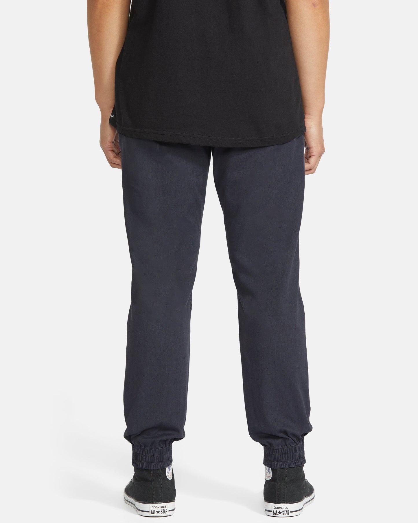 The lower half of a person faces away from the camera, wearing Volcom Men's Frickin Slim Joggers crafted with REPREVE recycled fibers, paired with a black t-shirt and black sneakers with white soles, set against a plain white background.