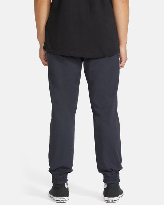 The lower half of a person faces away from the camera, wearing Volcom Men's Frickin Slim Joggers crafted with REPREVE recycled fibers, paired with a black t-shirt and black sneakers with white soles, set against a plain white background.