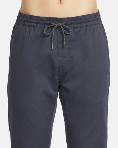 A close-up shows a person wearing Volcom Men's Frickin Slim Joggers in dark gray, featuring an elastic waistband with front drawstring and made with REPREVE recycled fibers.