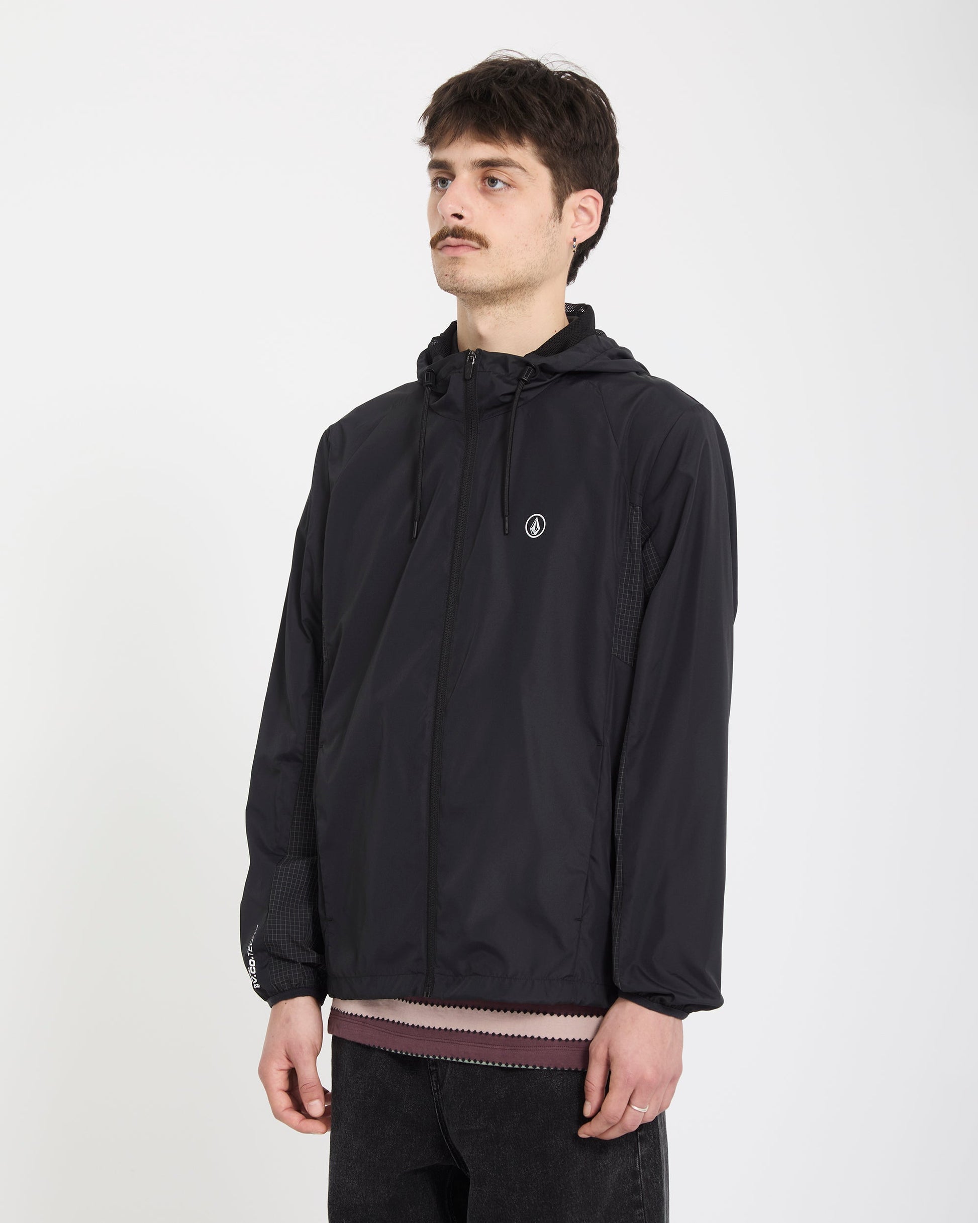 A young man with short brown hair and a mustache wears the Volcom Men's Wavern Jacket, featuring a small white logo on the chest, over a pink shirt and black pants, standing against a plain white background.