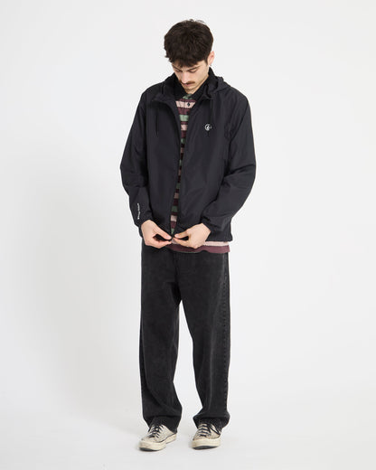 A person stands against a white background wearing the Volcom Men's Wavern Jacket over a striped shirt, with loose black pants and white sneakers. They look down while adjusting the front of the jacket.