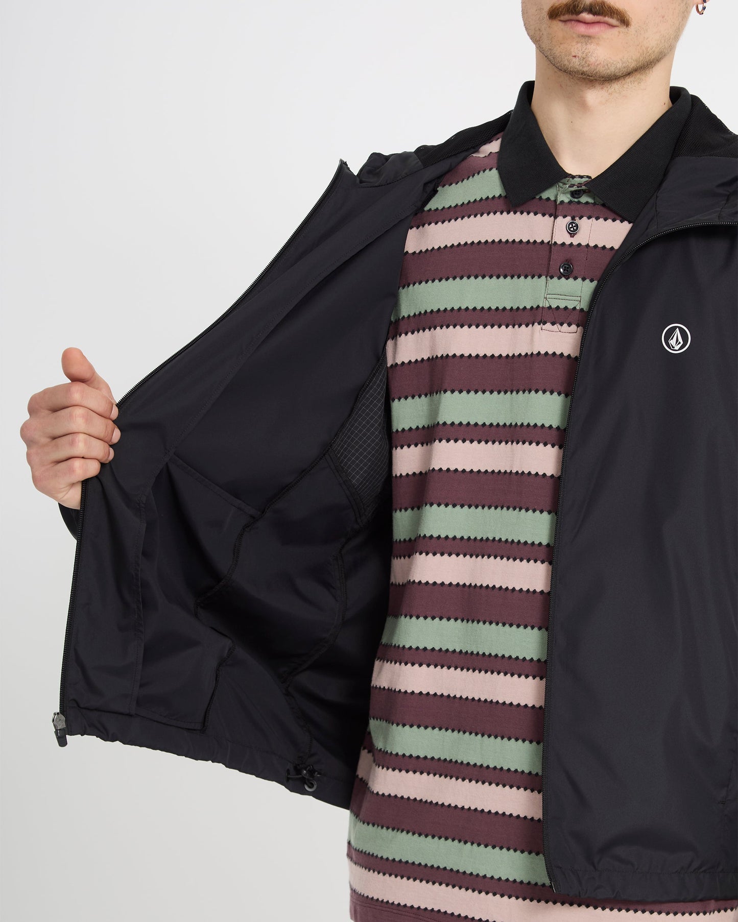 A man in a Volcom Men's Wavern Jacket opens it to show the inner pocket, revealing a striped polo shirt with green, pink, and maroon zigzag patterns. His face is partially visible, displaying a mustache.