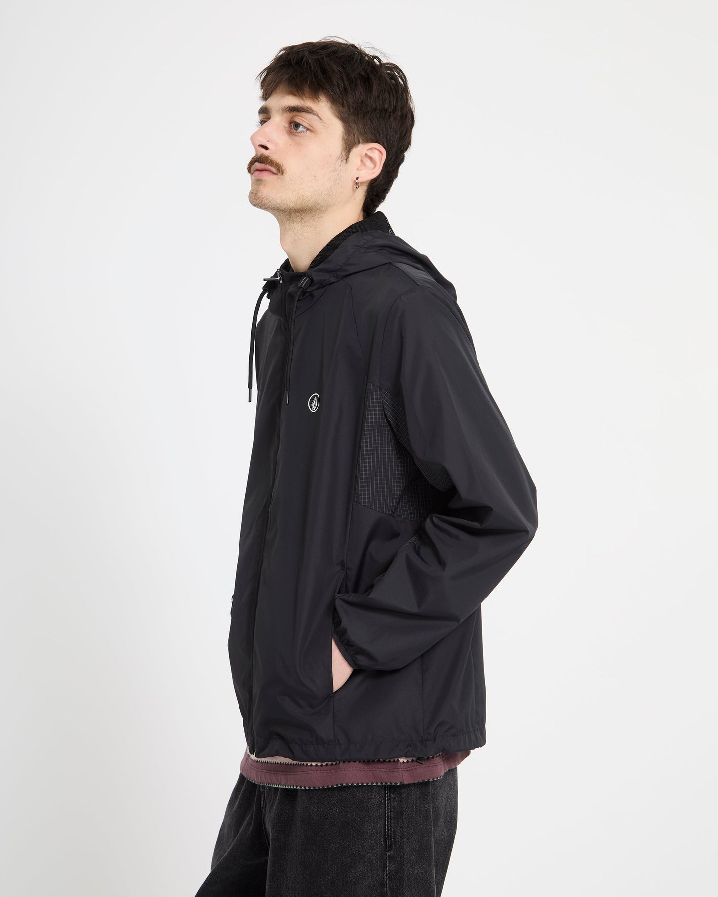 A young man with dark hair and a mustache wears the Volcom Men's Wavern Jacket, styled over a maroon shirt and dark jeans, standing sideways against a plain white background.