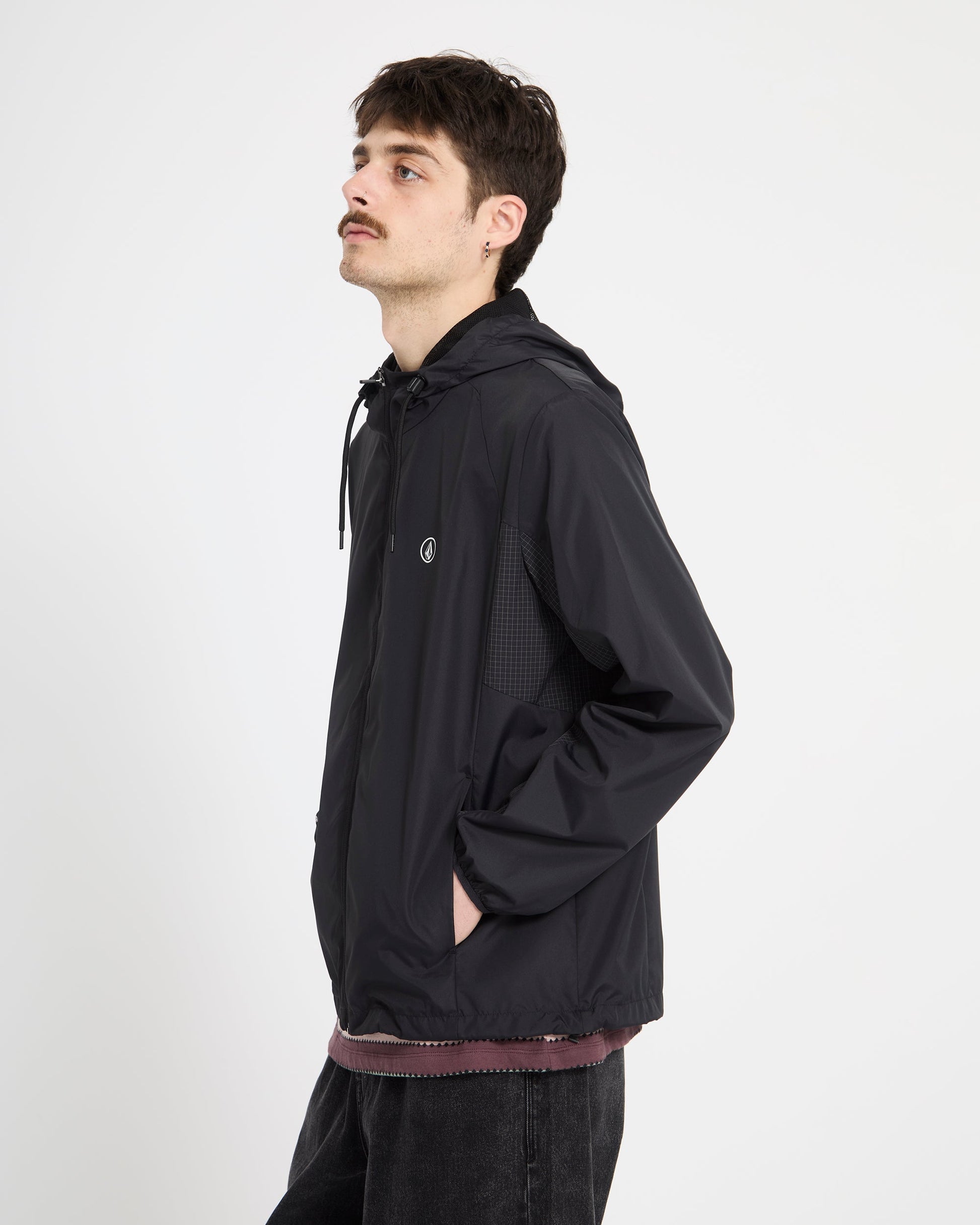 A young man with dark hair and a mustache wears the Volcom Men's Wavern Jacket, styled over a maroon shirt and dark jeans, standing sideways against a plain white background.