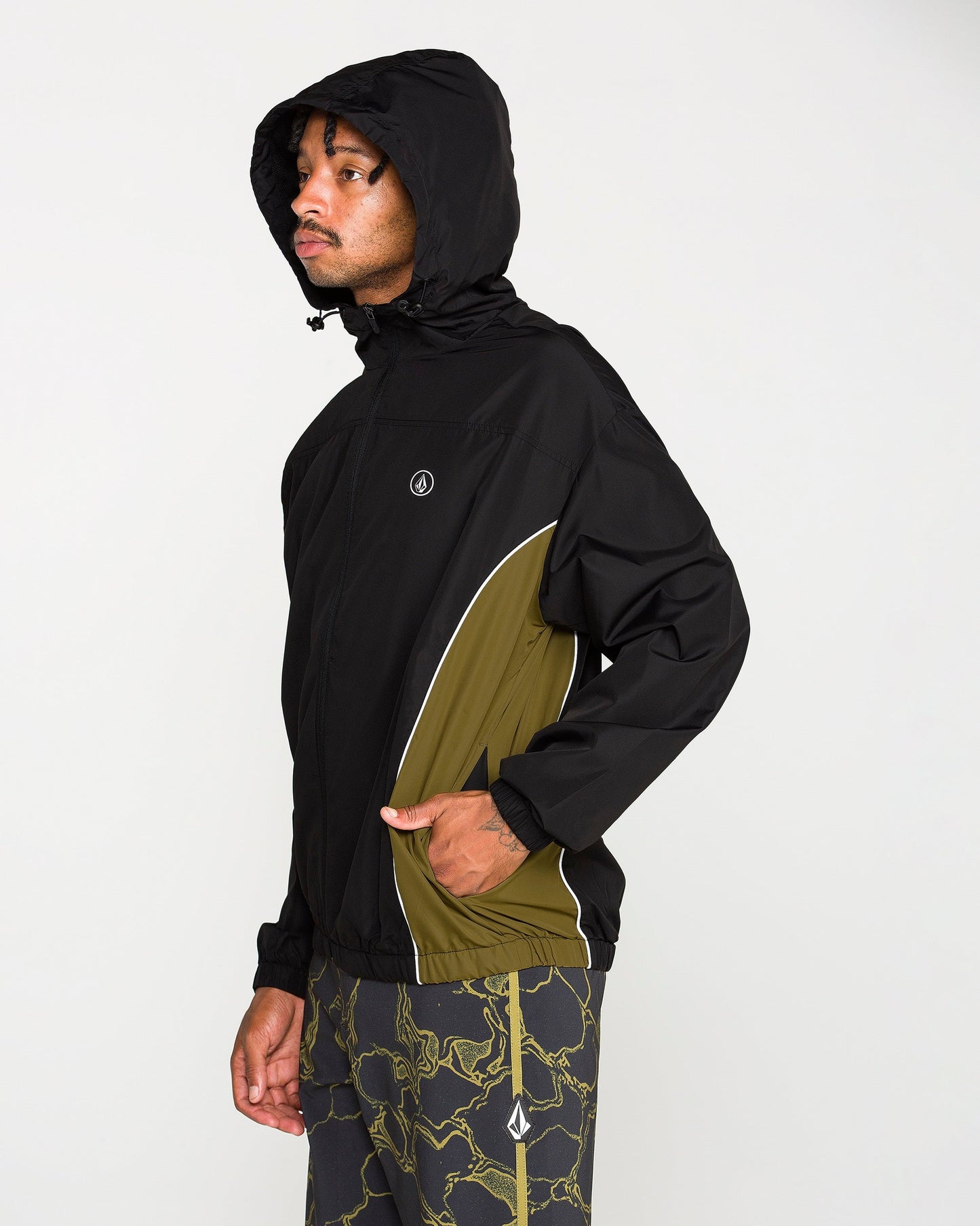 A man wearing the Volcom Men's Wavestone Jacket in black with olive green accents and a mesh-lined hood is paired with matching pants featuring a green lightning pattern, standing facing left against a plain white background.