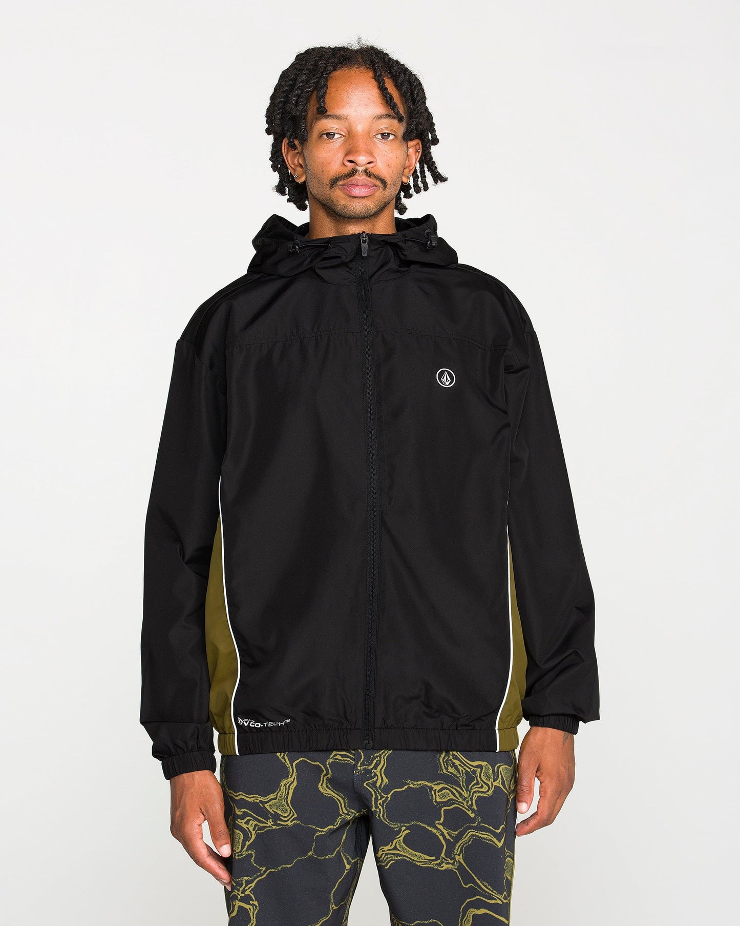 A person stands facing forward against a plain white background, wearing the Volcom Men's Wavestone Jacket in black, featuring a mesh-lined hood and white piping, paired with patterned black pants.