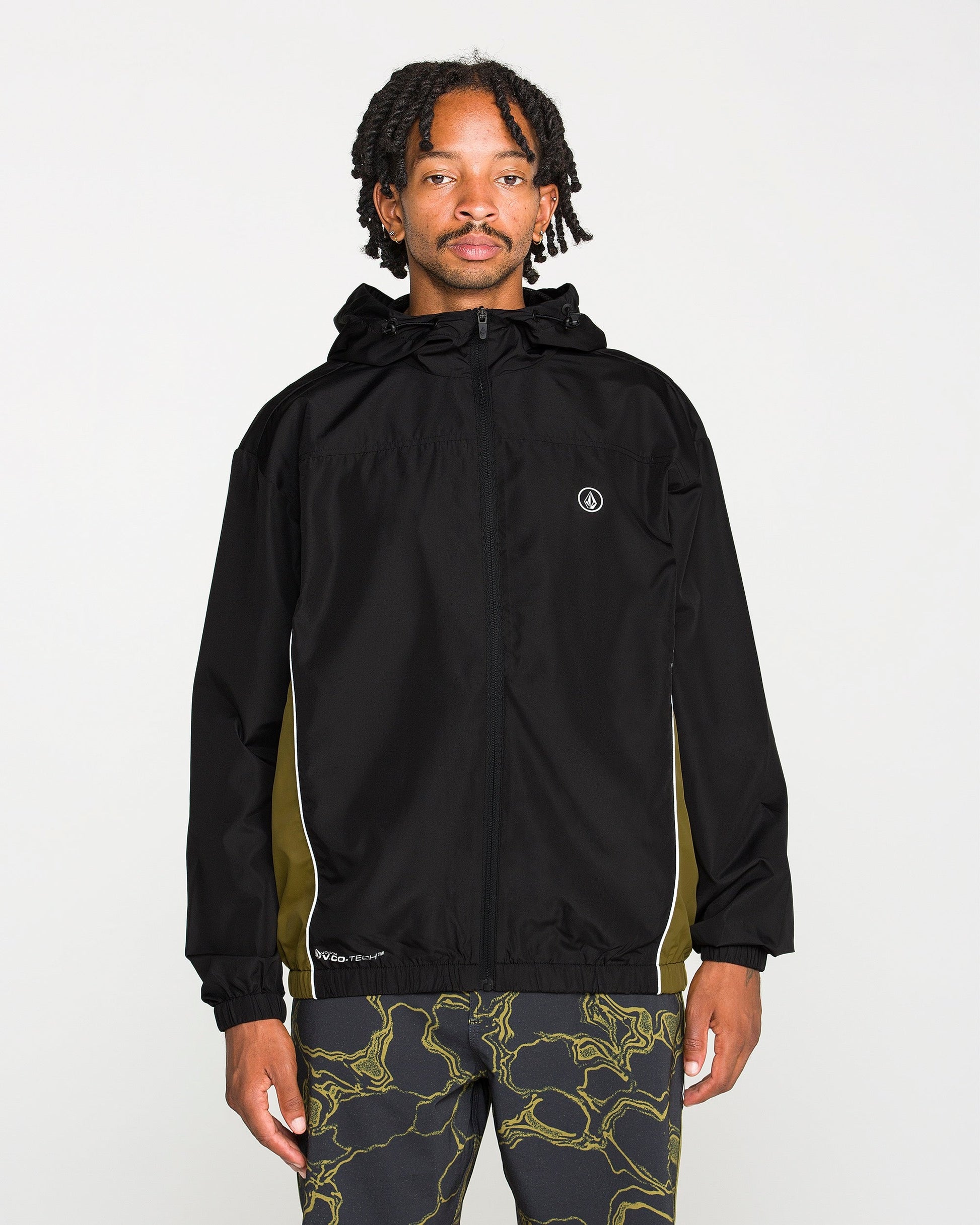 A person stands facing forward against a plain white background, wearing the Volcom Men's Wavestone Jacket in black, featuring a mesh-lined hood and white piping, paired with patterned black pants.