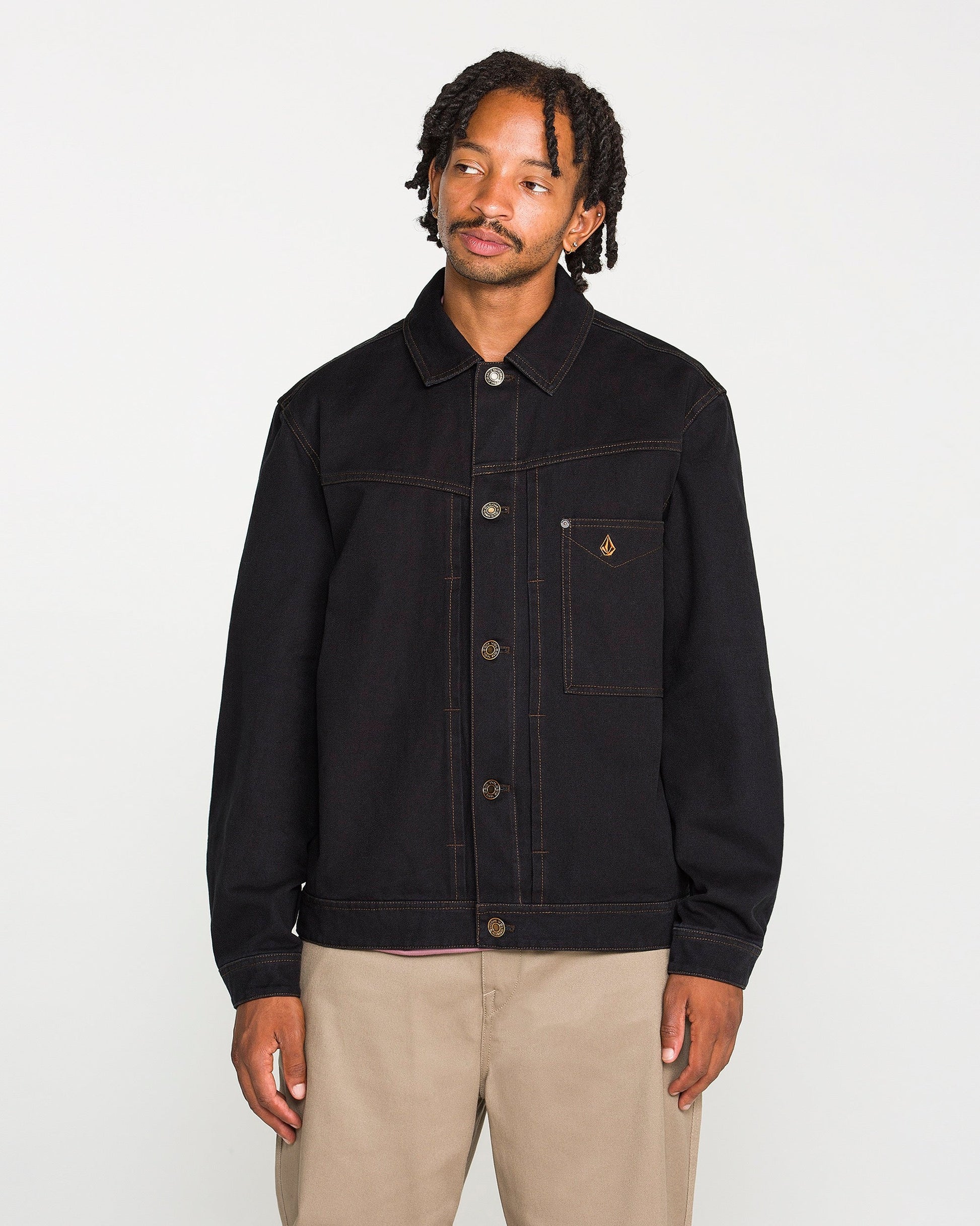 A person with short dreadlocks wears the Volcom Men's Slakers Jacket, a black lightweight piece featuring visible stitching and a small chest pocket, paired with beige pants while standing against a plain white background.