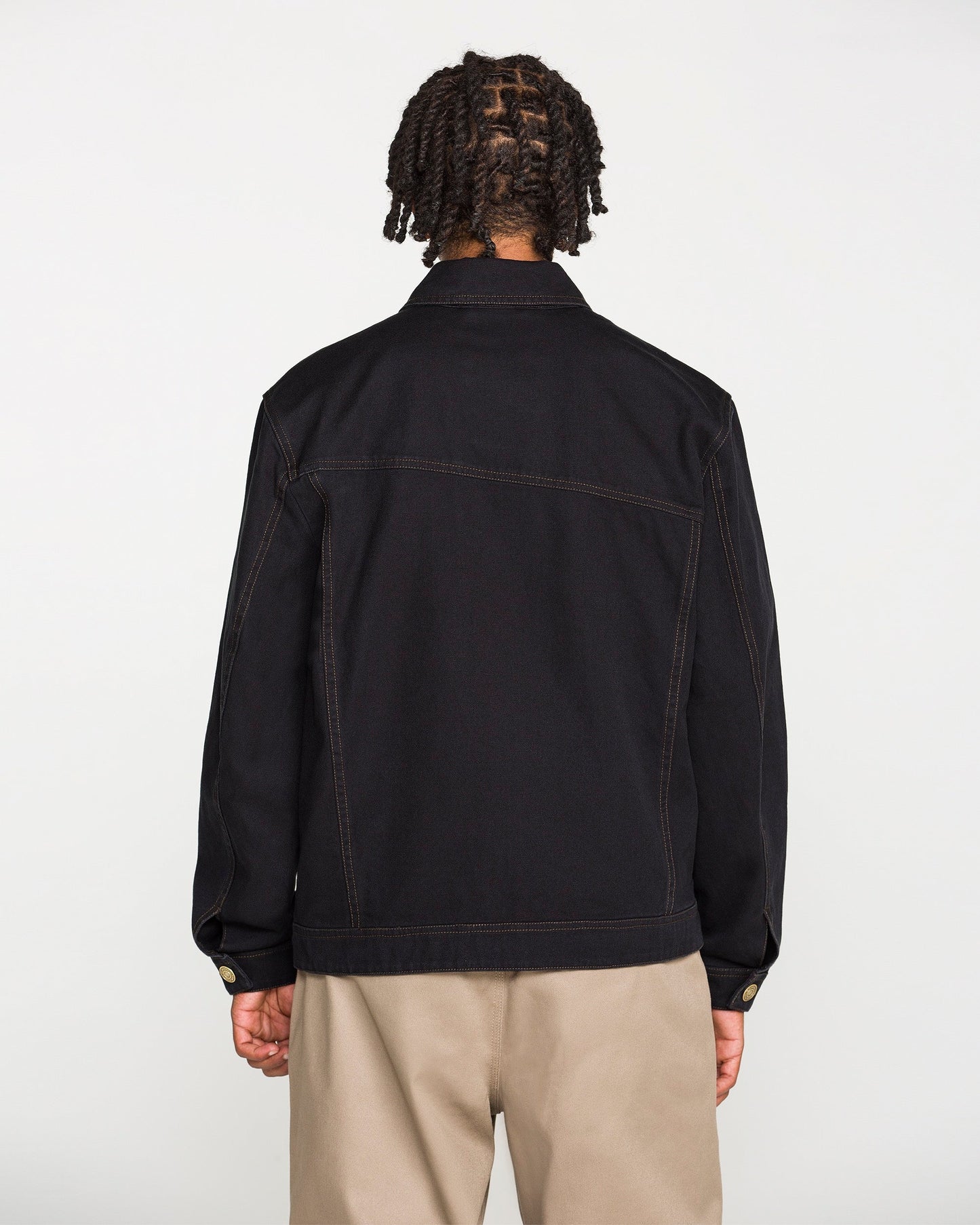 A person with short, twisted braids is shown from the back, wearing the Volcom Men's Slakers Jacket in black cotton canvas with visible seam lines and tan pants against a plain white background.