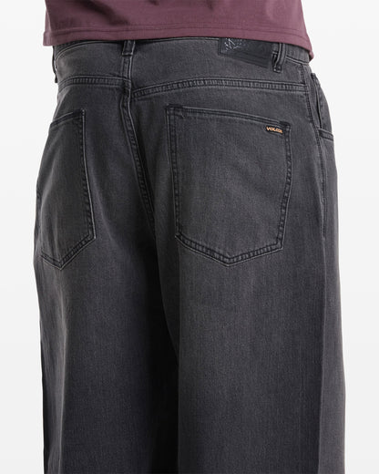 Close-up of the back of a person wearing Volcom Men's Chillow Denim in dark gray, featuring a relaxed fit, two back pockets, and a small Volcom logo tag above the right pocket, paired with a dark purple shirt.