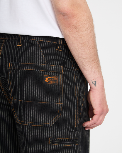 A person wears Volcom Men's Dialed Work Pants—black pinstripe with orange stitching and a brand patch on the back pocket. Their right arm, featuring a small wrist tattoo, is visible against a plain background.