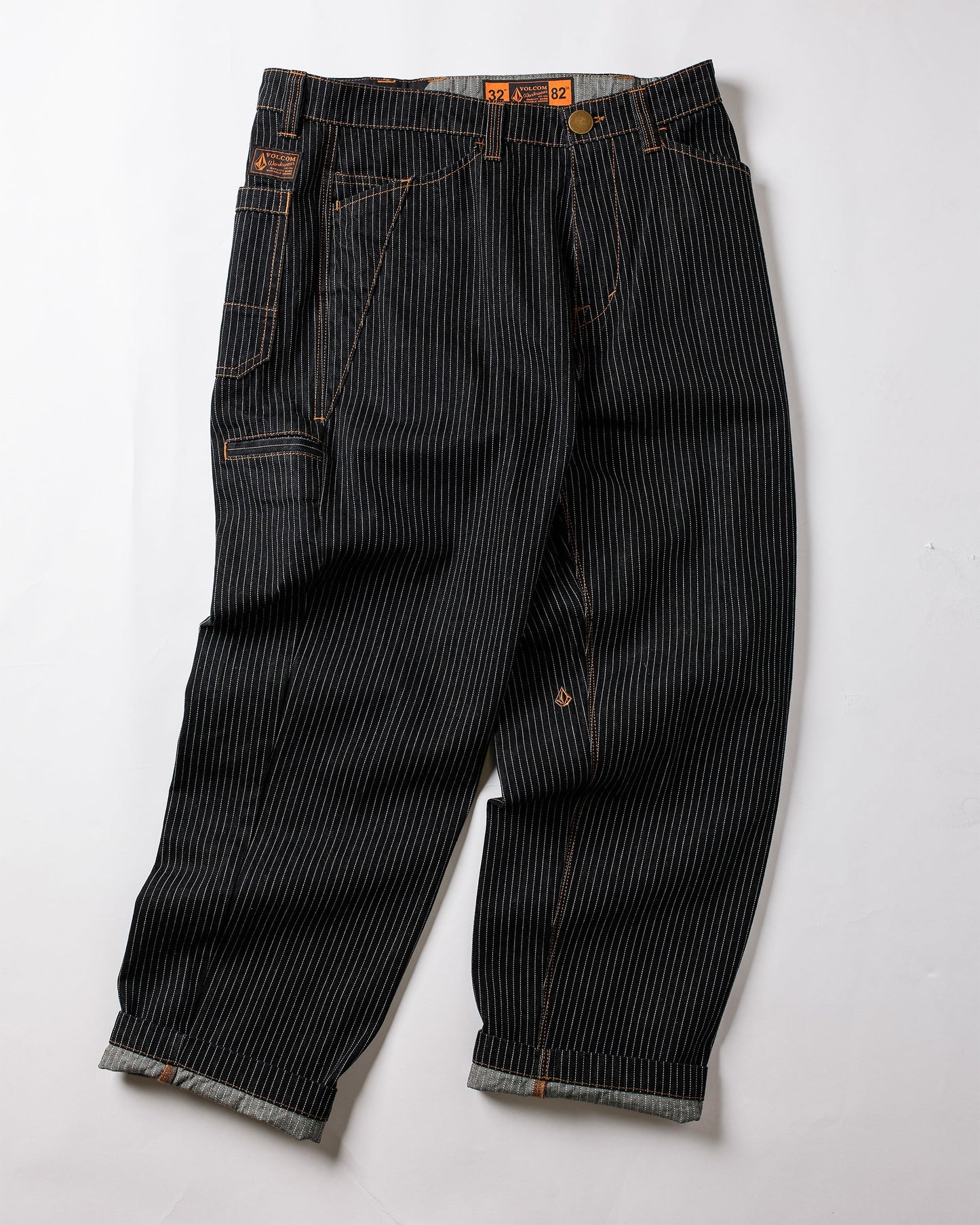 The Volcom Men's Dialed Work Pants feature black denim with white pinstripes, orange stitching, rolled cuffs, multiple pockets, and a metal waist button—combining durability and style for perfect workwear.