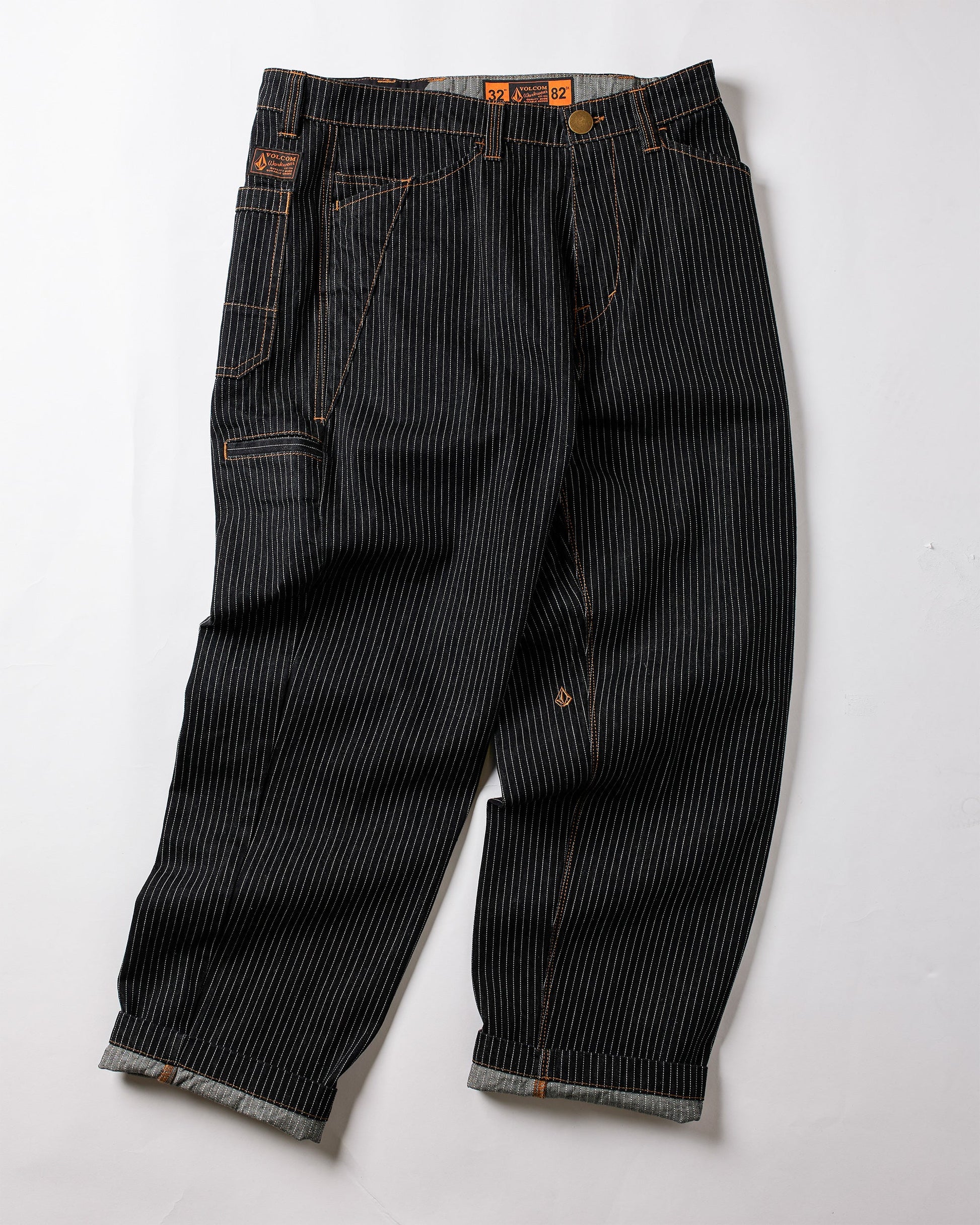 The Volcom Men's Dialed Work Pants feature black denim with white pinstripes, orange stitching, rolled cuffs, multiple pockets, and a metal waist button—combining durability and style for perfect workwear.