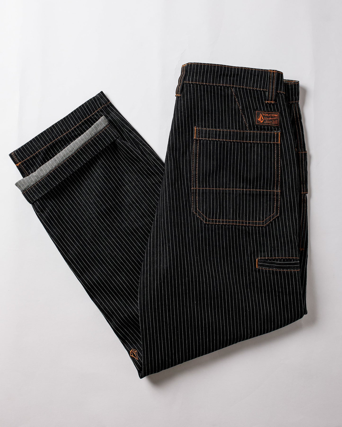 A pair of Volcom Men's Dialed Work Pants in black pinstripe with orange stitching, neatly folded on a white background. These durable canvas workwear pants have visible back pockets and a logo patch above the right pocket.