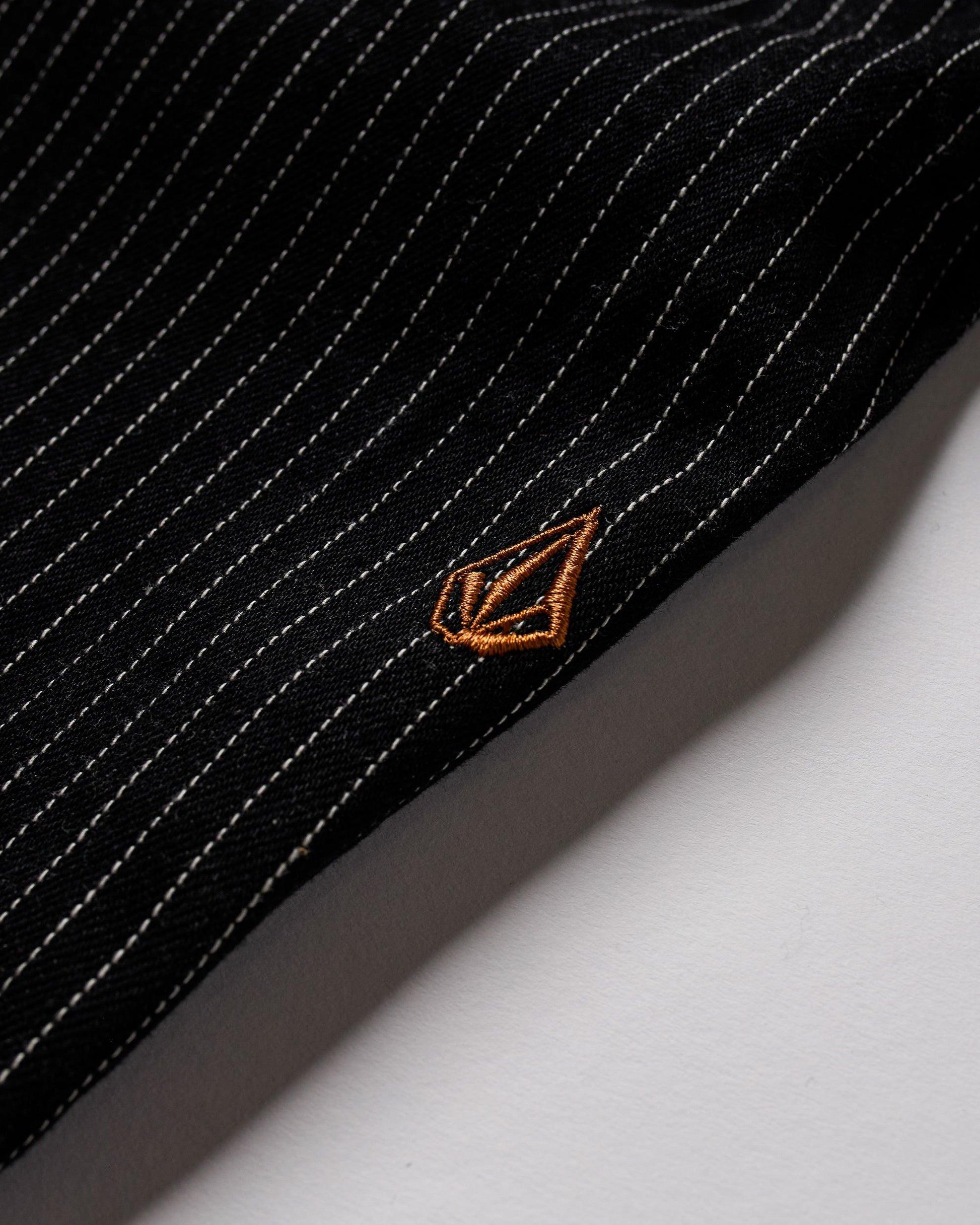 A close-up of durable Volcom Men's Dialed Work Pants fabric with thin white pinstripes and a small golden geometric embroidered logo near the edge, resting on a white surface—ideal for sturdy workwear.