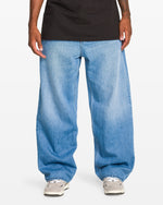 A person wears Volcom Men's Chillow Denim Vs jeans, paired with white sneakers and a black t-shirt, showing only their torso and legs against a plain white background.