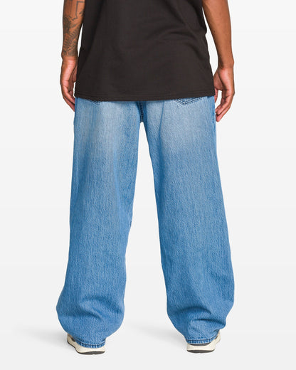 A person stands facing away, wearing a black T-shirt, Volcom Men's Chillow Denim Vs jeans with a baggy fit, and white sneakers. Their arms rest at their sides, revealing part of a tattoo on one forearm.