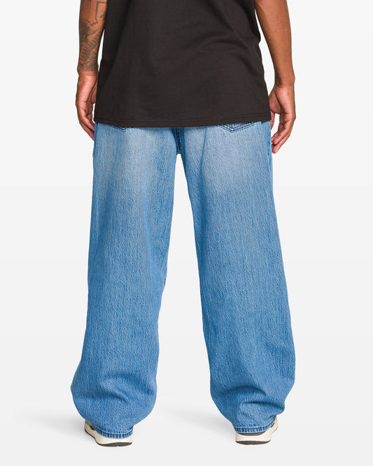A person stands facing away, wearing a black T-shirt, Volcom Men's Chillow Denim Vs jeans with a baggy fit, and white sneakers. Their arms rest at their sides, revealing part of a tattoo on one forearm.
