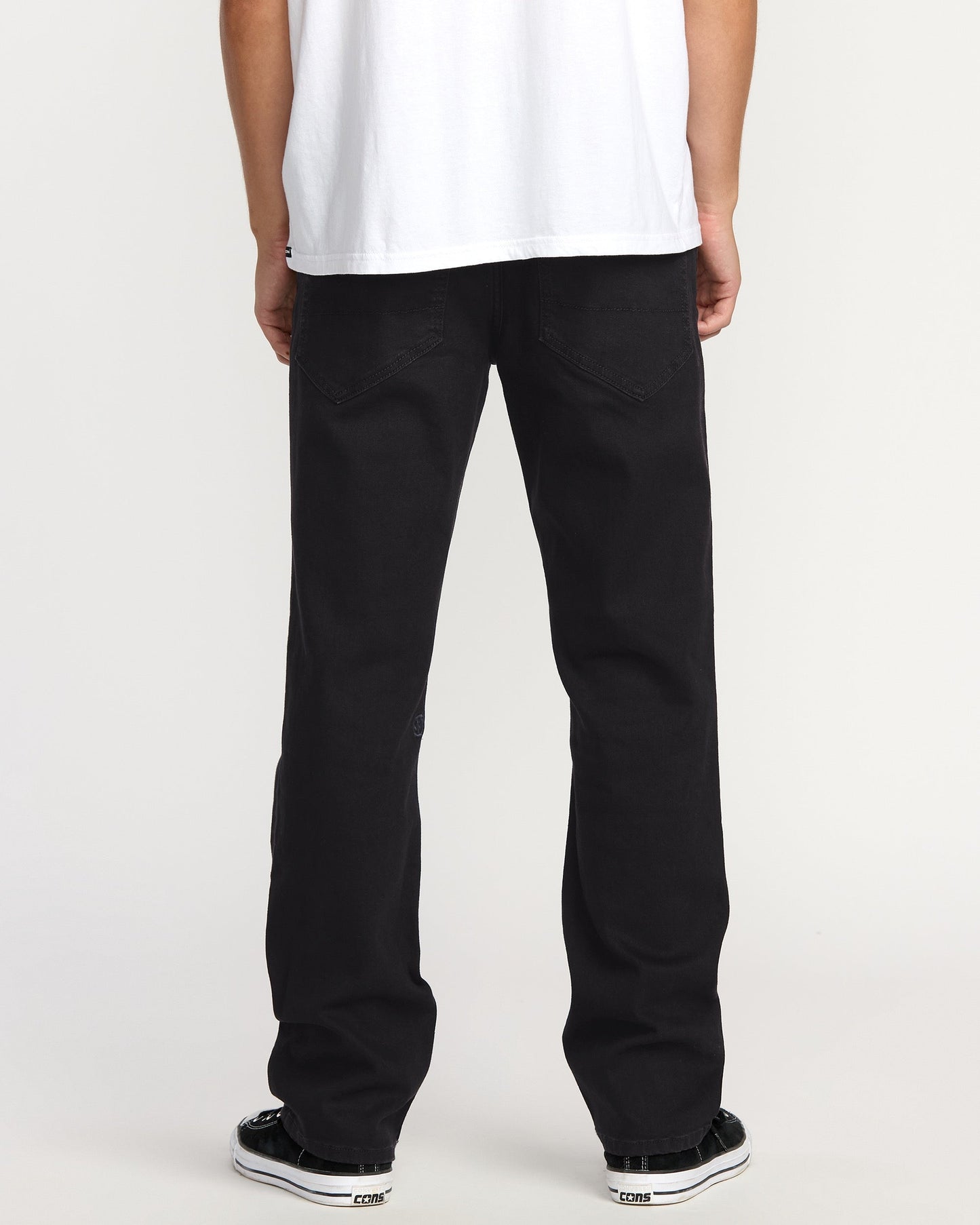 A person faces away, wearing a white T-shirt, black-and-white sneakers, and Volcom Men's Volcom Entertainment Pennywise Denim Pants, against a plain light backdrop.