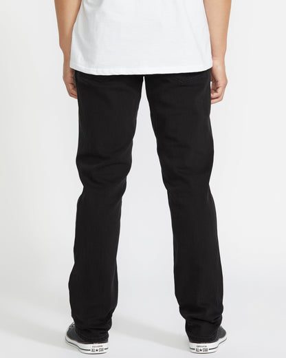 A person faces away from the camera, wearing a white shirt, Volcom Vorta Denim black stretch straight leg jeans, and black-and-white sneakers against a plain white background.