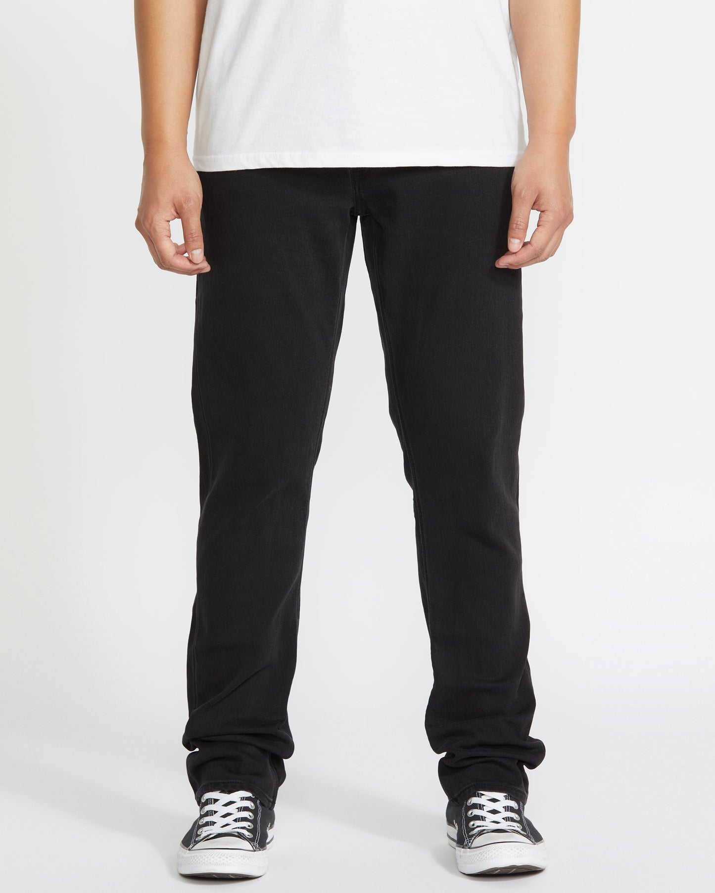 A person stands facing forward, wearing a white T-shirt, Volcom Vorta Denim black jeans, and black-and-white sneakers against a plain white background. The image is cropped from the shoulders down.