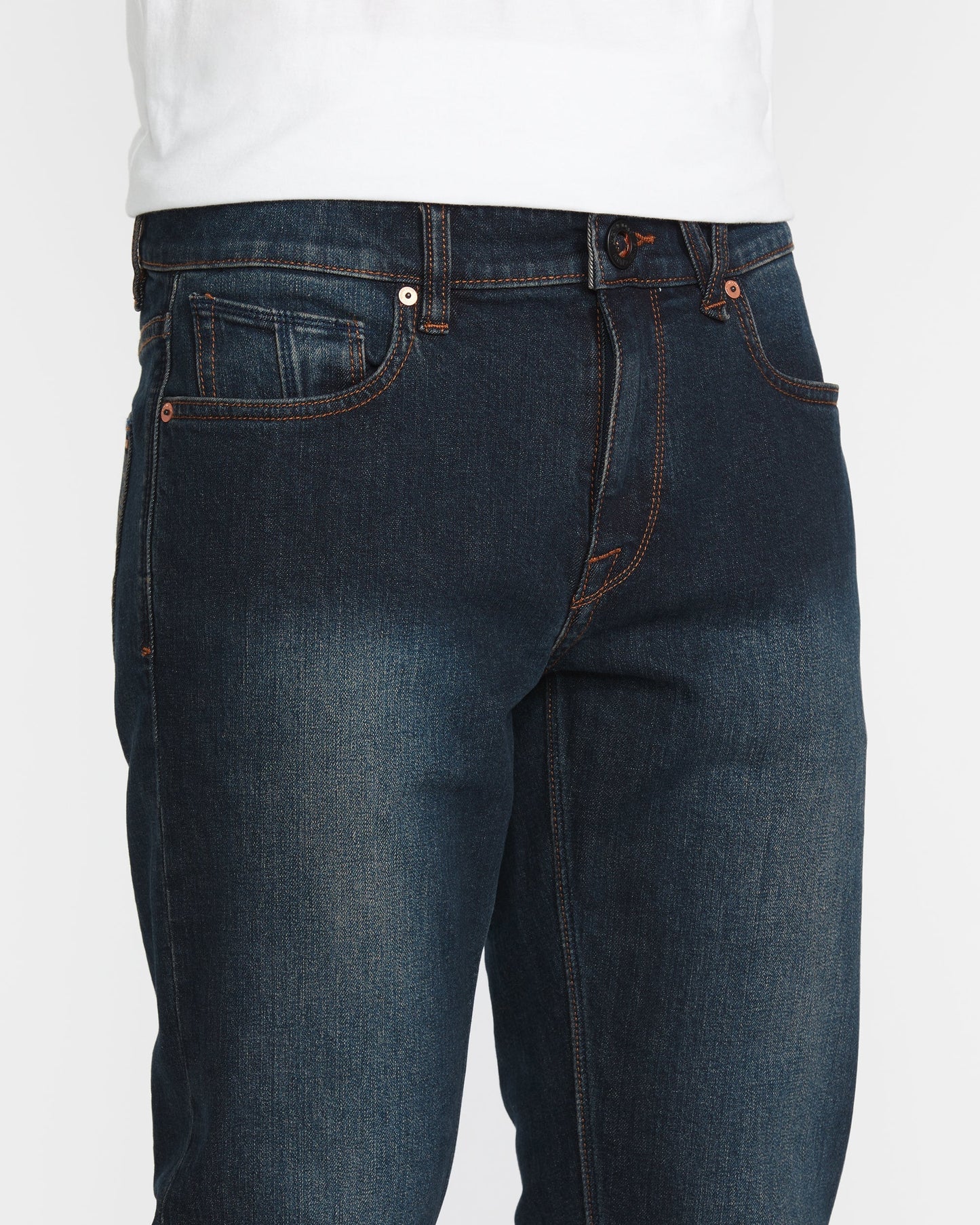 A person wearing Volcom Men's Vorta Denim jeans in dark blue with orange stitching and a white shirt. The image highlights the waist, upper legs, front pockets, and button closure.