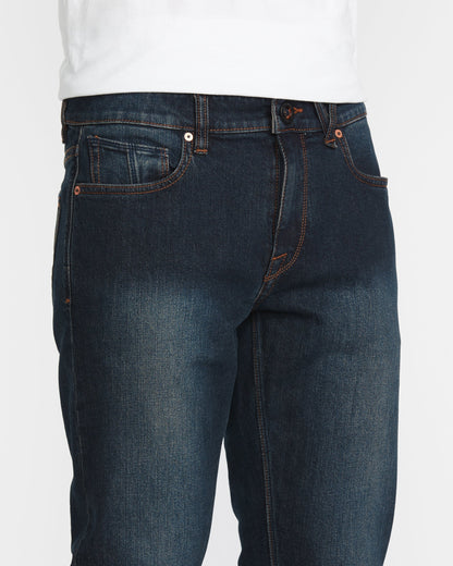 A person wearing Volcom Men's Vorta Denim jeans in dark blue with orange stitching and a white shirt. The image highlights the waist, upper legs, front pockets, and button closure.