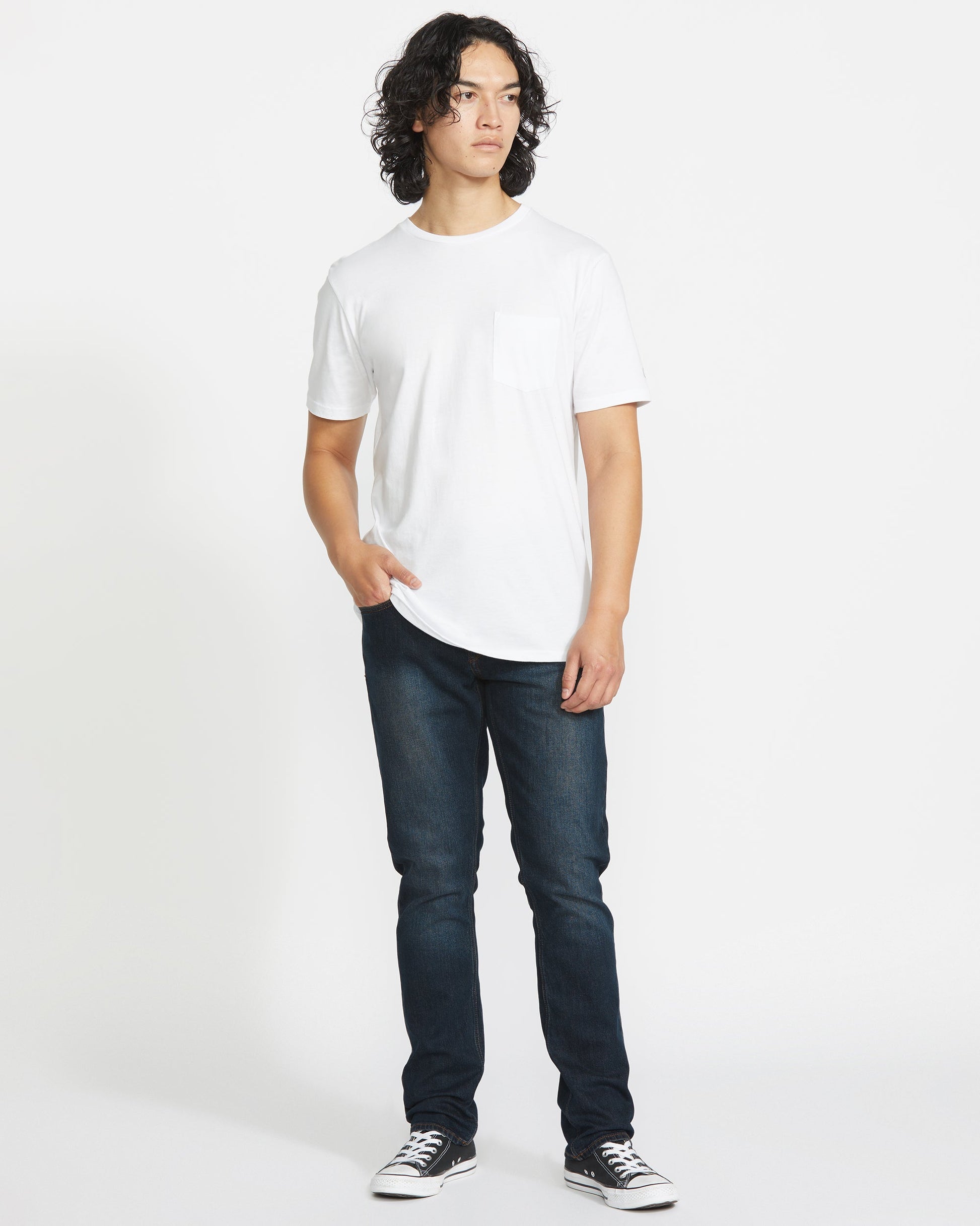 A person with curly hair stands against a white background, wearing a white short-sleeve t-shirt, Volcom Men's Vorta Denim jeans in dark blue, and black-and-white sneakers, with one hand in their pocket.