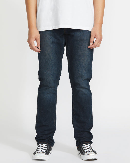 Wearing Volcom's Men's Vorta Denim jeans, a person stands against a plain white background. The image is cropped from the shoulders down and also shows a white t-shirt and black-and-white sneakers.