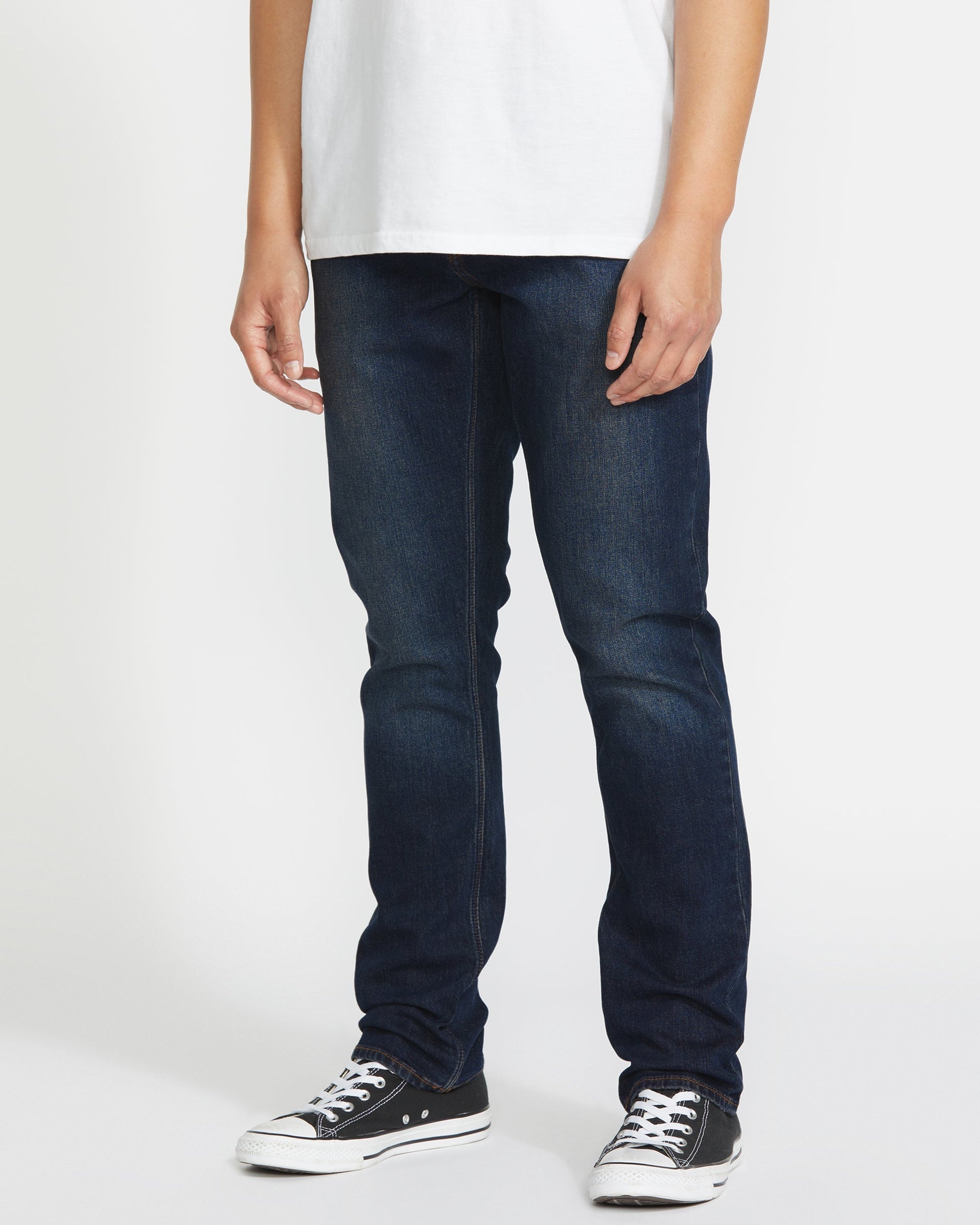 A person stands against a plain white background, showing only their lower half while wearing Volcom Men's Vorta Denim jeans, a plain white t-shirt, and black low-top Converse sneakers.