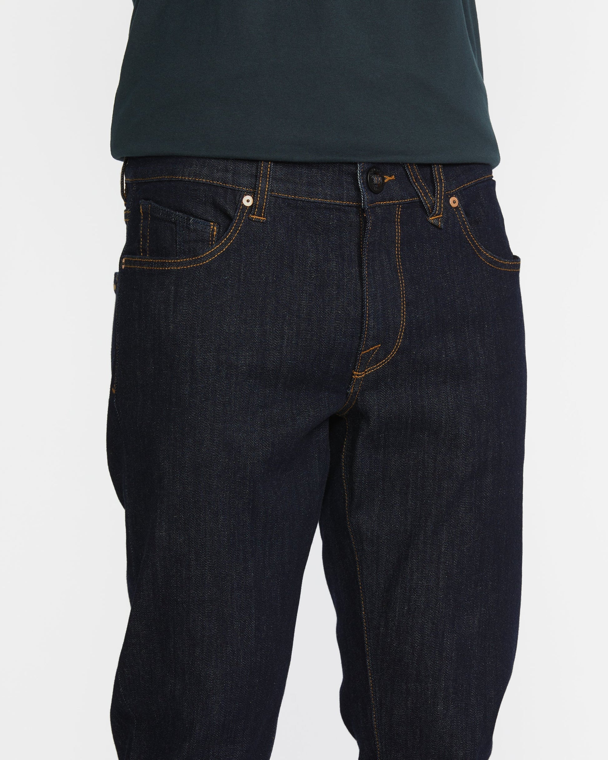 A person wears slim fit Men's Vorta Denim by Volcom with orange stitching and a dark green shirt. The photo focuses on the waist and upper legs against a white background, highlighting Water Aware details.