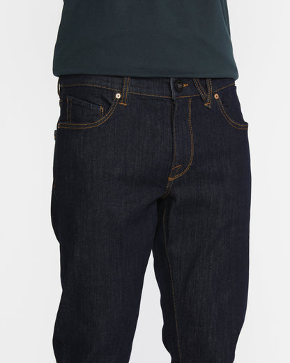 A person wears slim fit Men's Vorta Denim by Volcom with orange stitching and a dark green shirt. The photo focuses on the waist and upper legs against a white background, highlighting Water Aware details.
