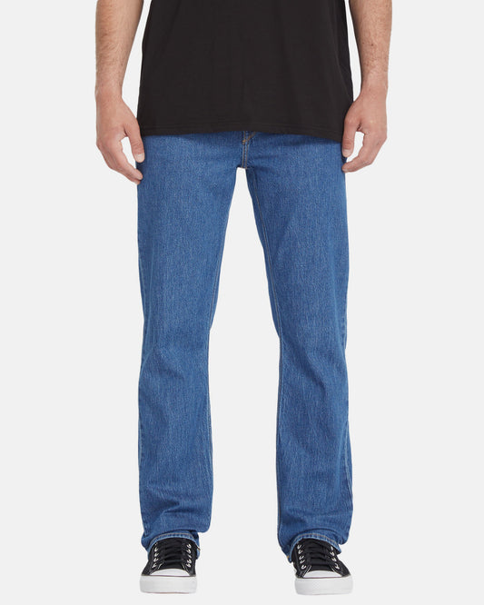 A person wearing a black t-shirt, Volcom Men's Solver Denim Pants in blue, and black-and-white sneakers, shown from the shoulders down against a plain white background.