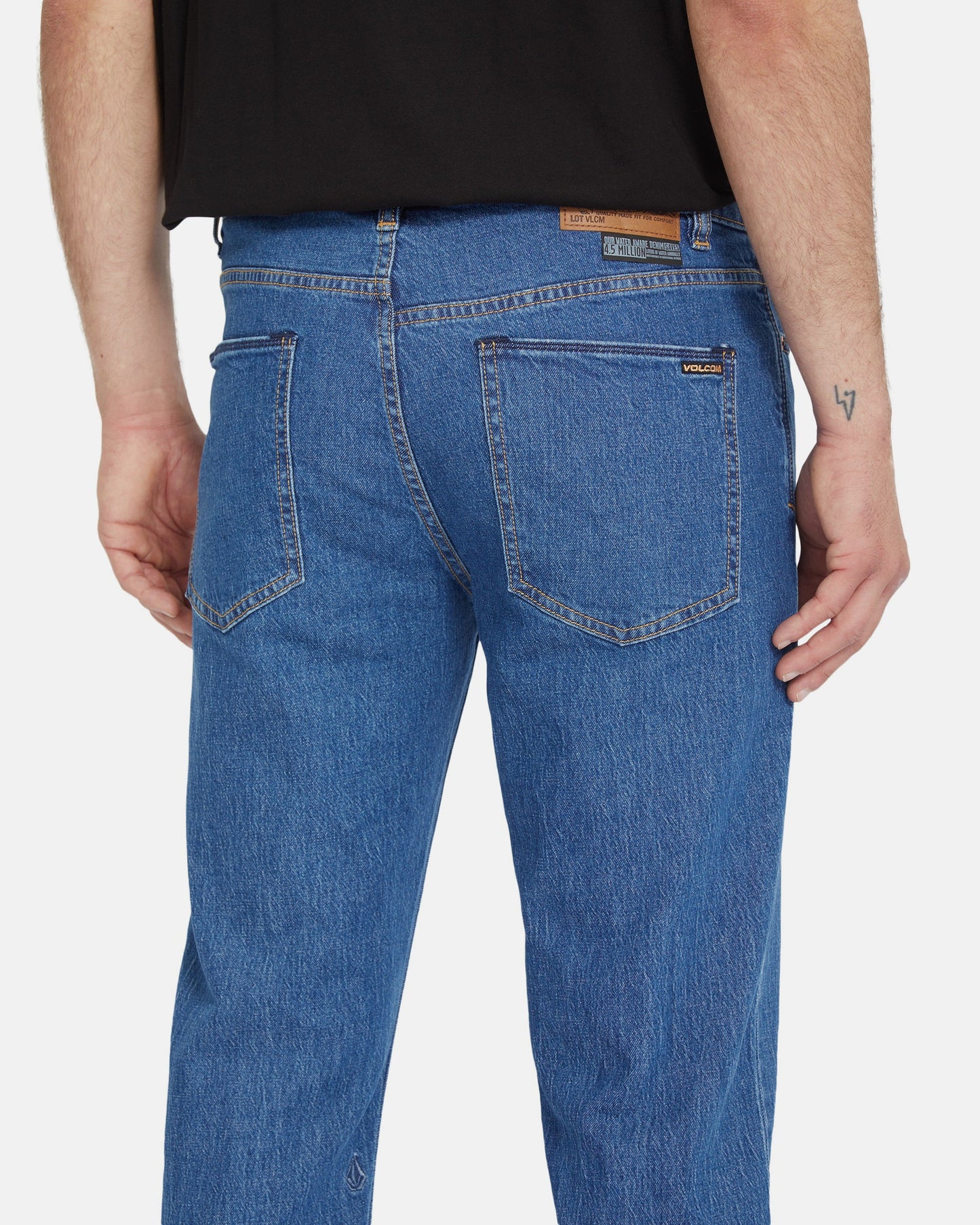 A person is shown from the back wearing Volcom Men's Solver Denim Pants in blue with two back pockets, a brown leather patch on the waistband, and a black shirt. A small tattoo appears on their right arm.