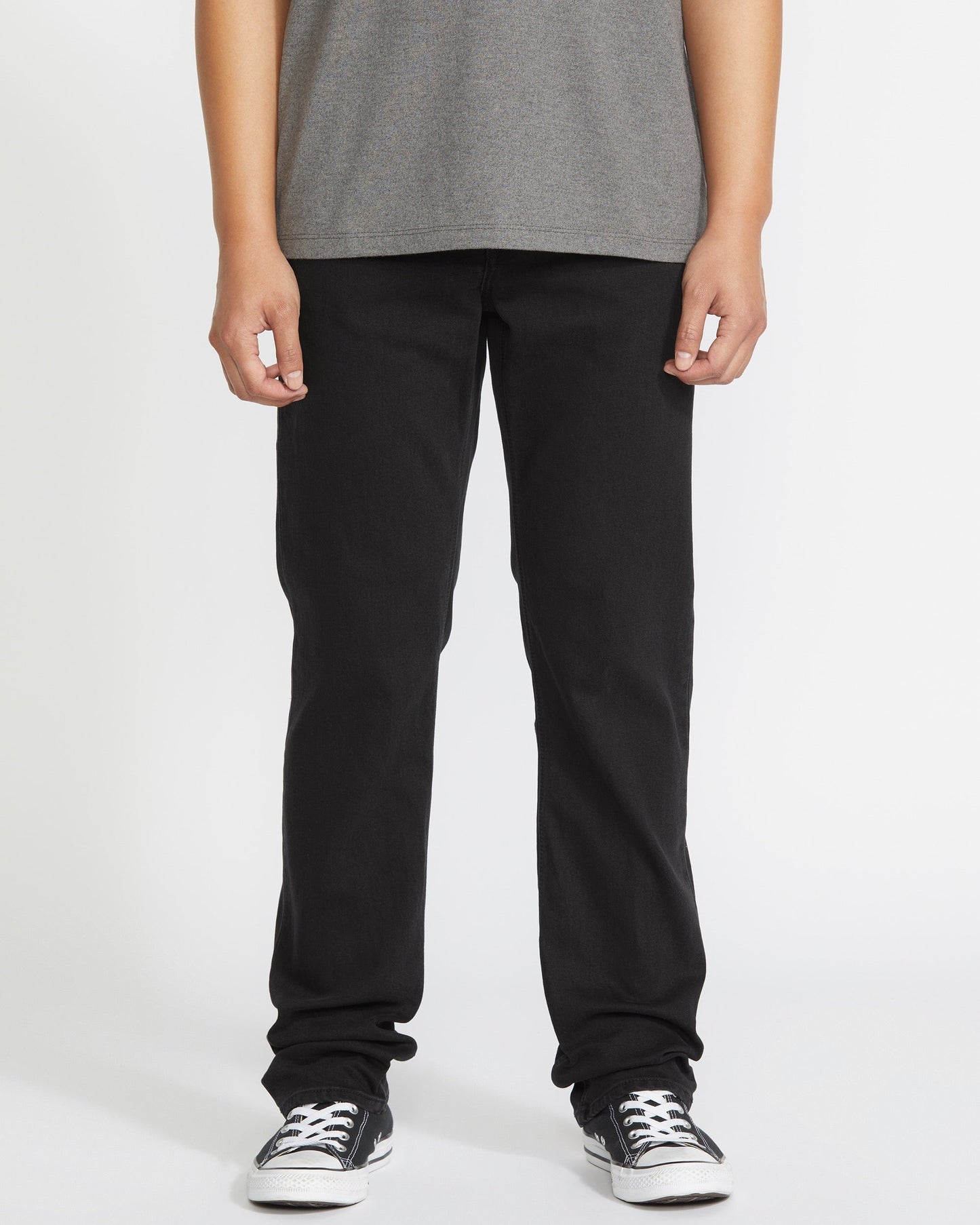 A person is seen from the waist down, wearing a gray t-shirt, Volcom Men's Solver Denim Pant in black, and black-and-white sneakers against a plain white background.