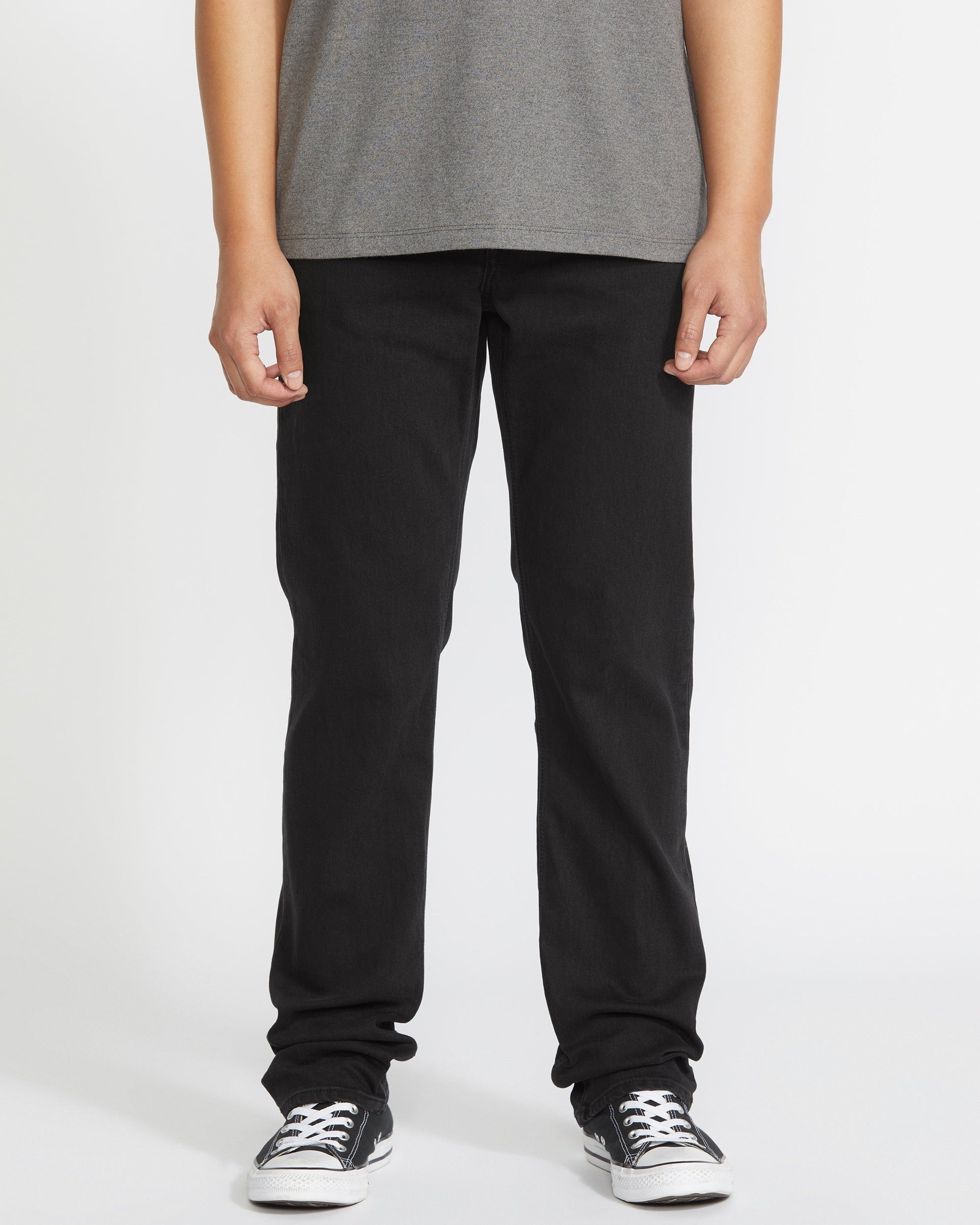 A person is seen from the waist down, wearing a gray t-shirt, Volcom Men's Solver Denim Pant in black, and black-and-white sneakers against a plain white background.